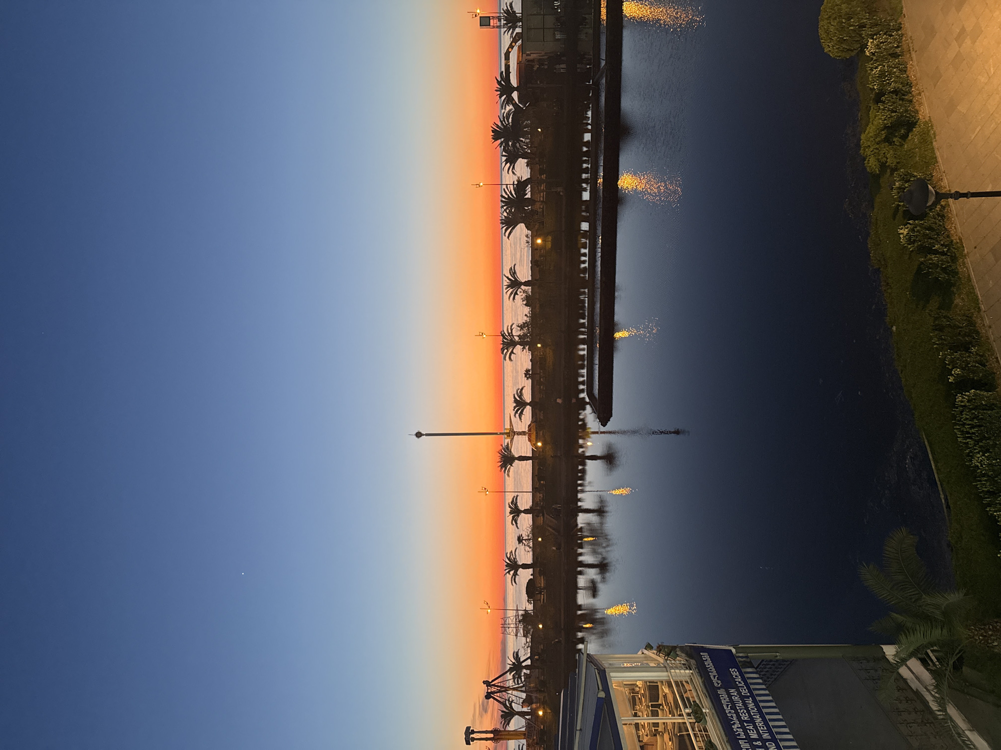The image shows a view in Batumi, Georgia, at dusk. The sky is a gradient of blue to orange at the horizon, where it meets the sea. A line of palm trees and lampposts lines the shore. There is a pier extending into the water. The water reflects the lights along the shore and the sky. On the right side of the image, there is a landscaped area with green bushes and a sidewalk. On the bottom, there is a building with a sign in Georgian and English, indicating it is a restaurant. The lighting suggests it is close to sunset or twilight.