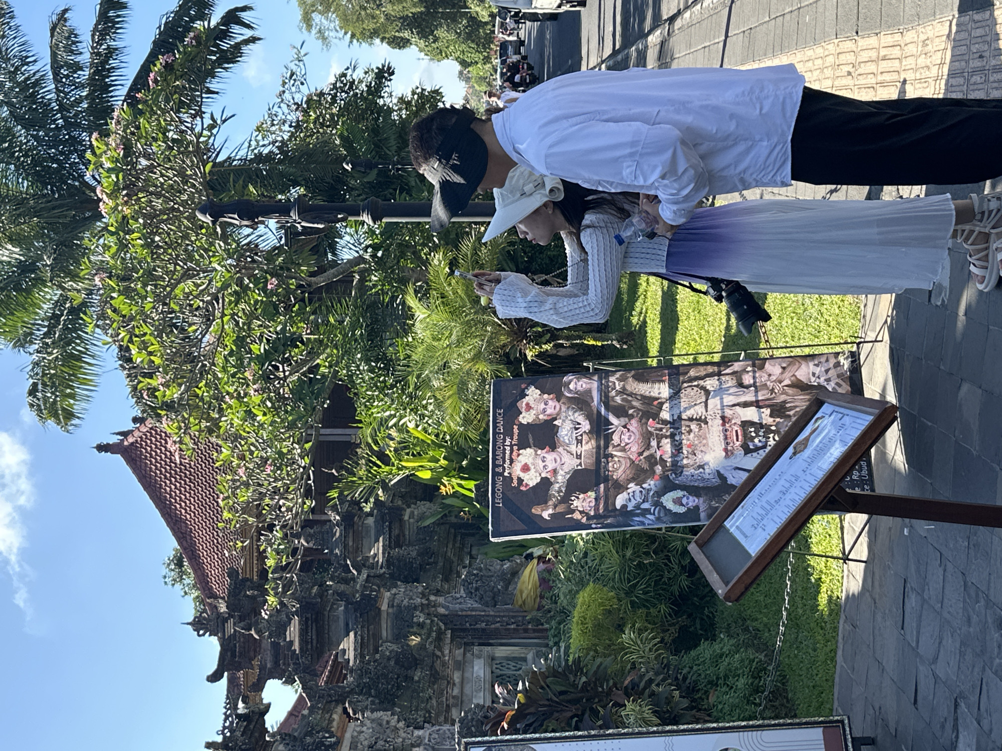 In the city of Ubud District, Indonesia, on a bright, sunny day, a couple stands on a paved sidewalk next to a street lined with vehicles and palm trees. The woman, wearing a white sunhat and a long white and purple dress, is taking a photo with her phone. A camera hangs from her neck. The man, in a white shirt and dark pants, stands beside her. In the background, a poster advertises "LEGONG & BARONG DANCE" performed by the "Sadha Budaya Troupe". Behind the poster, a traditional Balinese building with intricate stonework and a red tile roof is visible, surrounded by lush green foliage. A wooden easel displays a menu or program in front of the building.