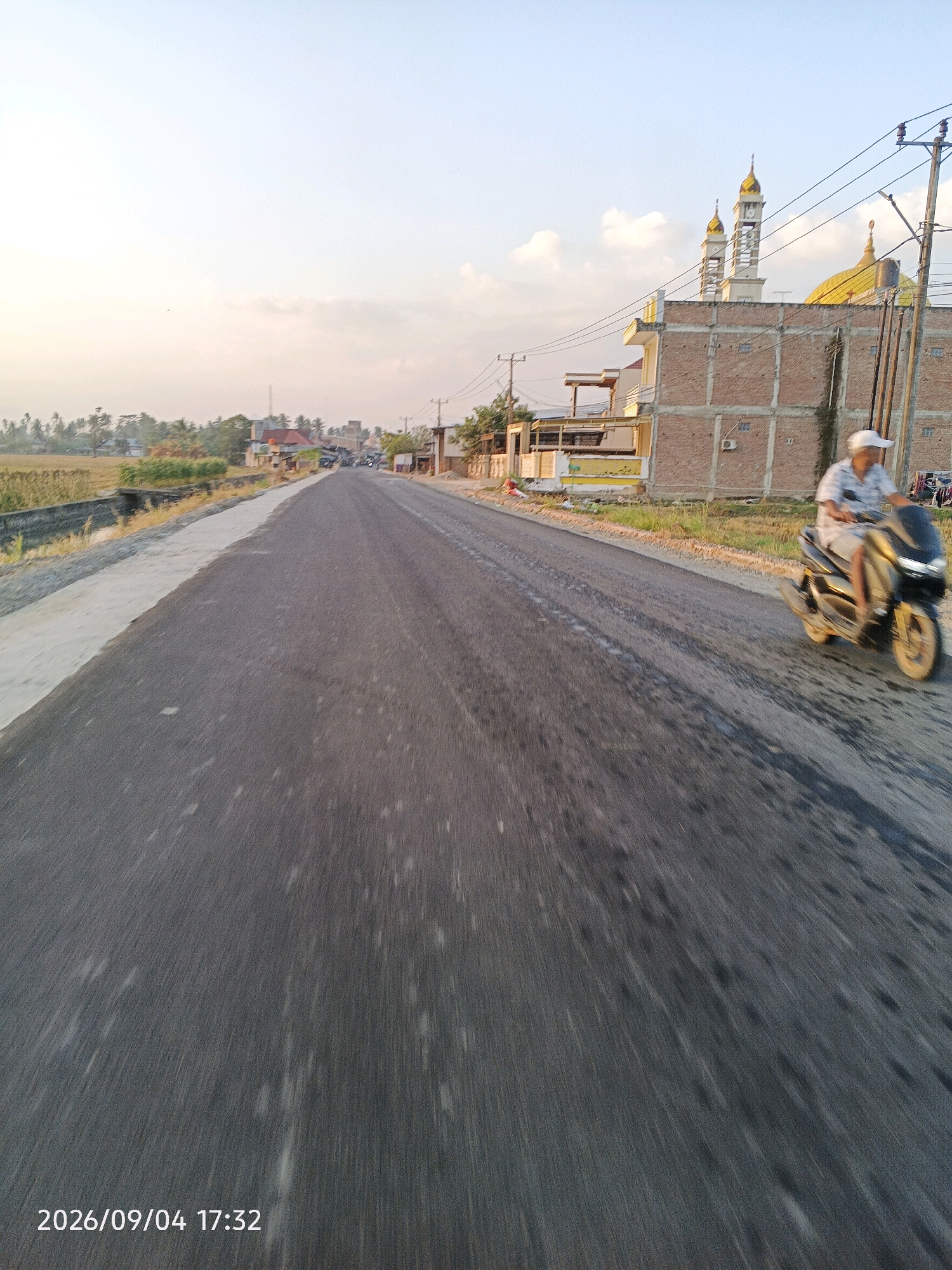 The image depicts a paved road in Bulu Cenrana, Indonesia, with a rural or suburban setting. The road is in the foreground, with a slight blur indicating motion. To the right of the road, a person is riding a motorcycle. Behind the motorcycle, there is a brick building with a golden-domed structure and minarets, likely a mosque. To the left of the road, there are agricultural fields and scattered buildings. The sky is bright and hazy, with some clouds, suggesting it is daytime, likely late afternoon given the soft lighting. The timestamp at the bottom of the image reads "2026/09/04 17:32".