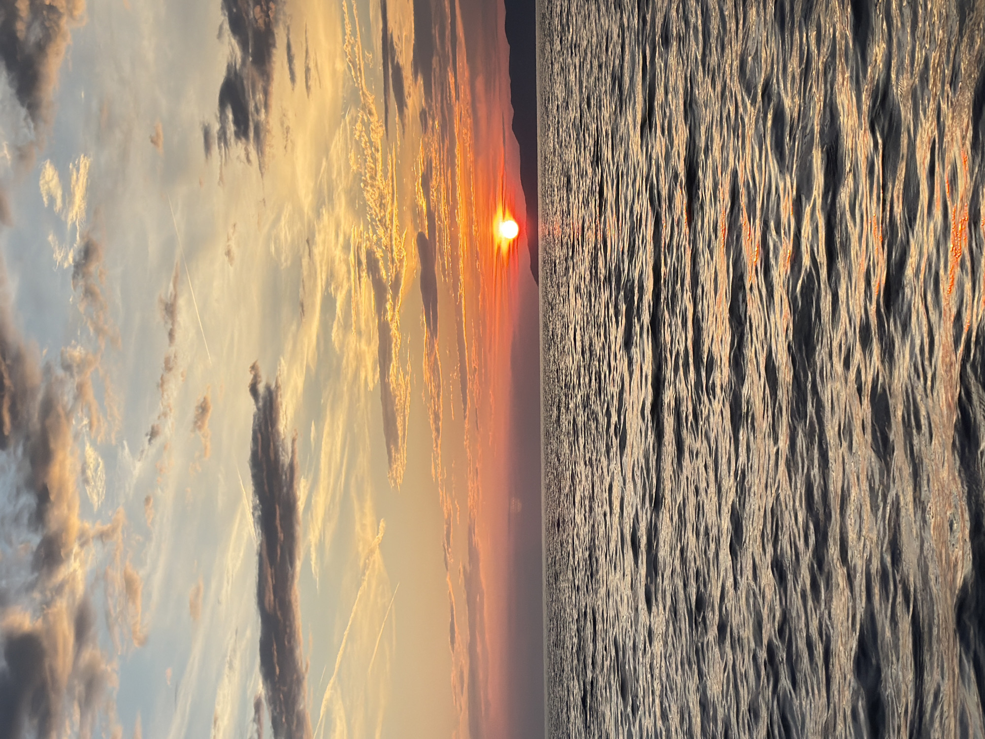 The image depicts a sunset over the Adriatic Sea, viewed from Petrovac na Moru, Montenegro. The sun, a bright orange orb, is partially obscured by a silhouetted mountain range in the distance. The sky is a vibrant gradient of orange, pink, and blue, with scattered clouds tinged with yellow and grey. The sea is calm, with gentle ripples reflecting the colors of the sunset. The water in the foreground is a deep, dark blue, transitioning to lighter hues closer to the horizon. The scene is devoid of any people, boats, or other man-made objects, emphasizing the natural beauty of the coastal landscape.