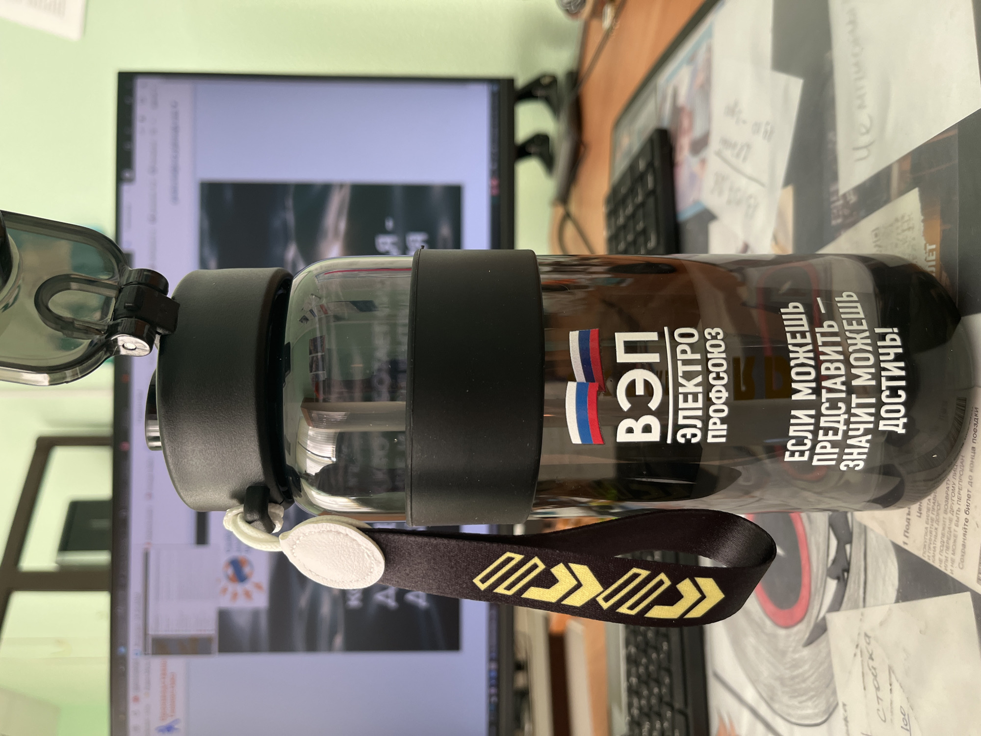 Here is a detailed description of the image:
**Subjects and Objects:**
* A translucent water bottle with a black lid, neck strap, and rubber grip.
* A black keyboard and monitor on a cluttered desk.
* Papers and objects are scattered around the desk.
**Text and Graphics:**
* The water bottle has Russian text that reads "ВЭП ЭЛЕКТРО ПРОФСОЮЗ".
* The bottle also says "ЕСЛИ МОЖЕШЬ ПРЕДСТАВИТЬ - ЗНАЧИТ МОЖЕШЬ ДОСТИЧЬ!" which translates to "If you can imagine it, you can achieve it!"
* The neck strap has yellow directional arrows on a black background.
* A piece of paper reads "Стойка".
**Environment:**
* The setting appears to be an office environment.
* The background includes a computer monitor and assorted office supplies.
**Location:**
* Chaikovsky, Russia.
* The acronym "ВЭП" (VEP) stands for "Всероссийский Электропрофсоюз", the All-Russian Electrical Workers' Union.
