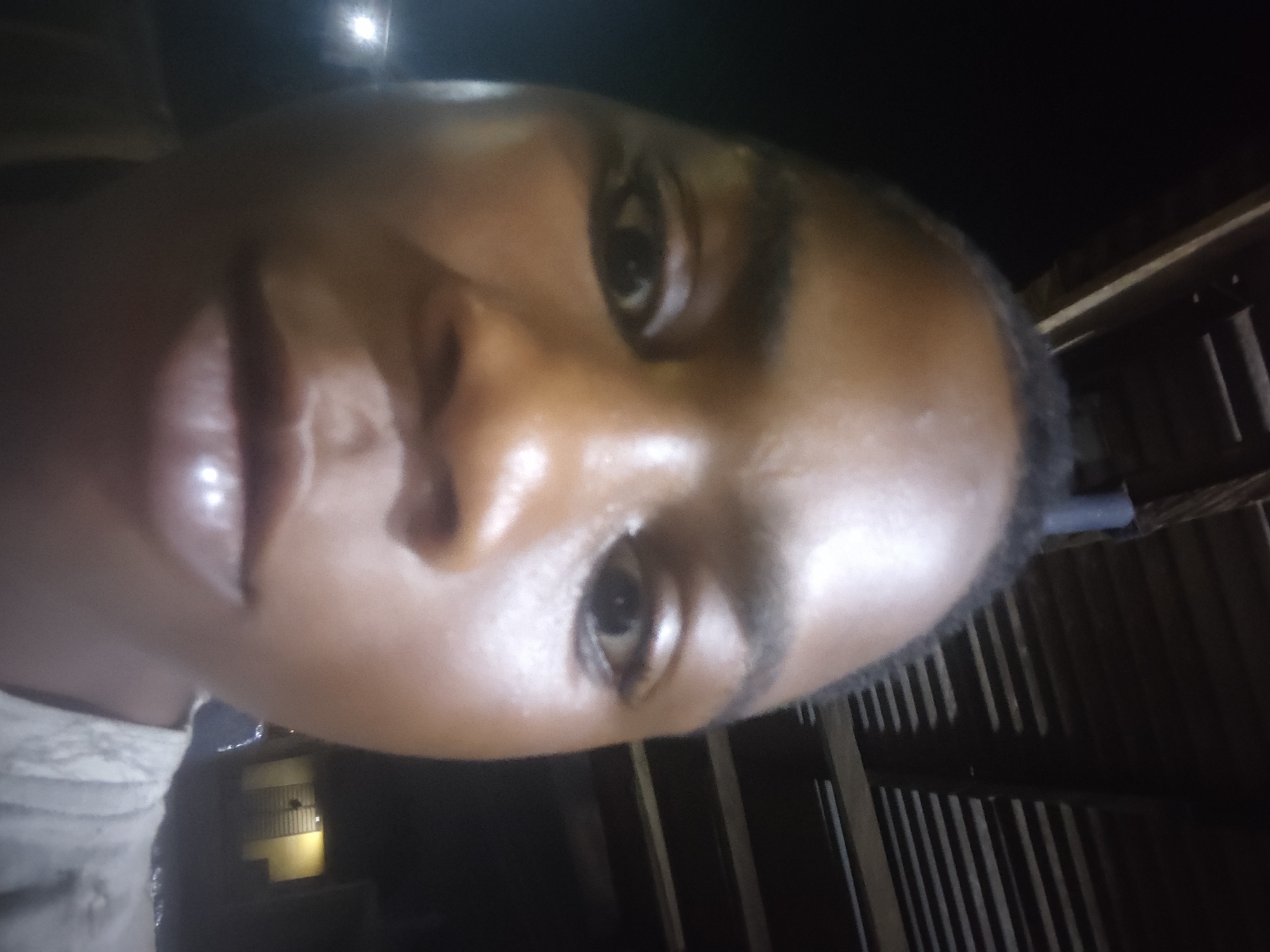 A close-up selfie of a Black person's face with dark eyes is the central subject of the image. The face is well-lit, highlighting smooth skin. To the right of the subject is a dark, possibly indoor space defined by what appears to be vertical wooden planks or slats. In the background, a faint glow emanates from a distance, hinting at an outdoor setting with some illumination. The image's composition is tilted, and it's difficult to establish the precise location within Ikorodu, Nigeria, without further context or recognizable landmarks.