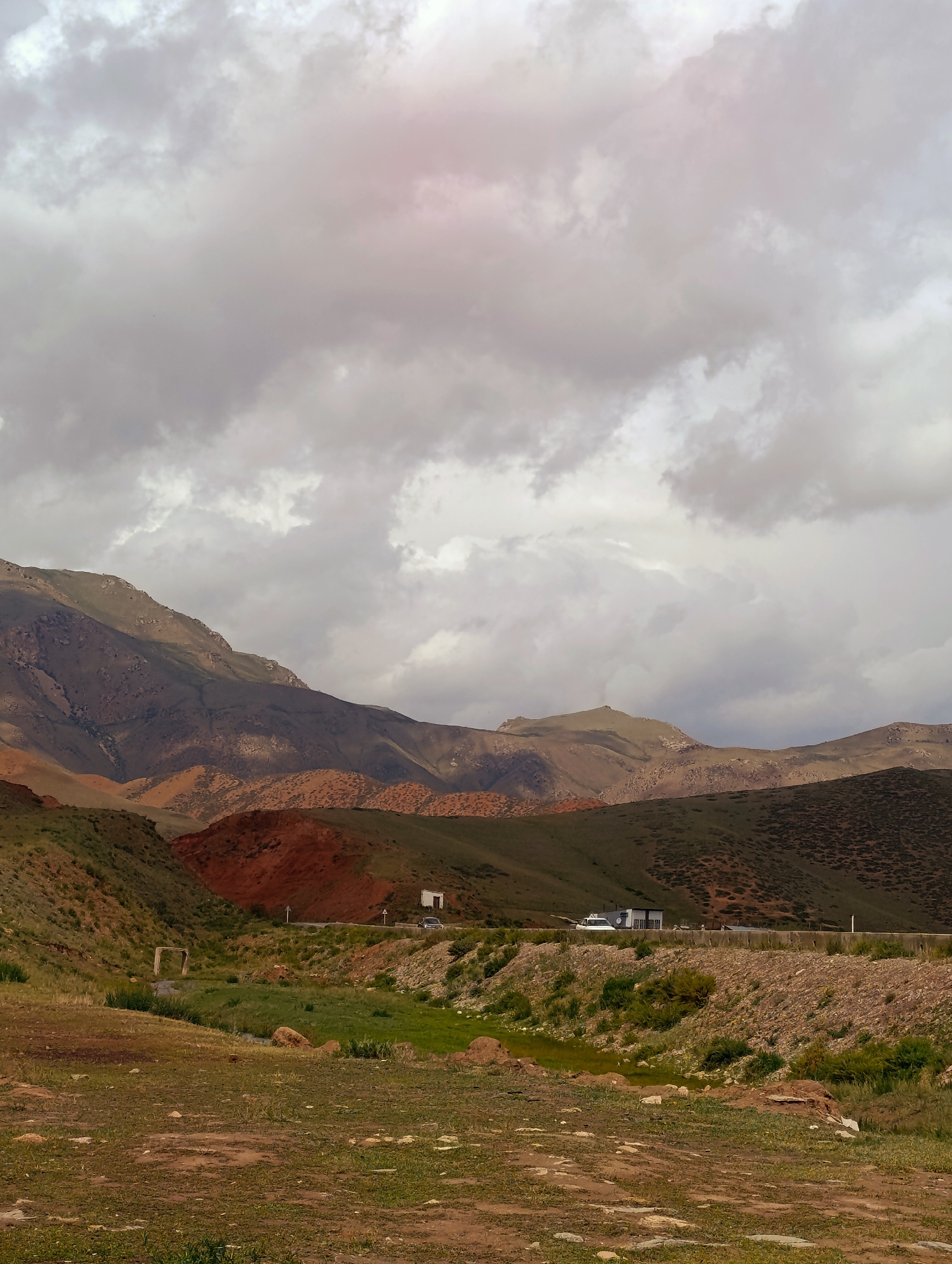 The image depicts a landscape in the vicinity of Ак Жар, Kyrgyzstan, characterized by rolling hills and mountains under a cloudy sky. In the foreground, dry, brown earth with sparse green vegetation is visible. A small stream or water channel meanders through a patch of lush green grass in the middle ground. Beyond the stream, a road runs horizontally across the frame, with a white vehicle and a small building or structure situated alongside it. The hills in the middle and background display a variety of colors, including green, brown, and reddish-orange, with some areas appearing barren and others covered in low-lying vegetation. The distant mountains are a muted brown and green, fading into the hazy sky. The clouds are heavy and grey, with hints of pinkish light suggesting the possibility of sunrise or sunset. No people are visible in the image.
