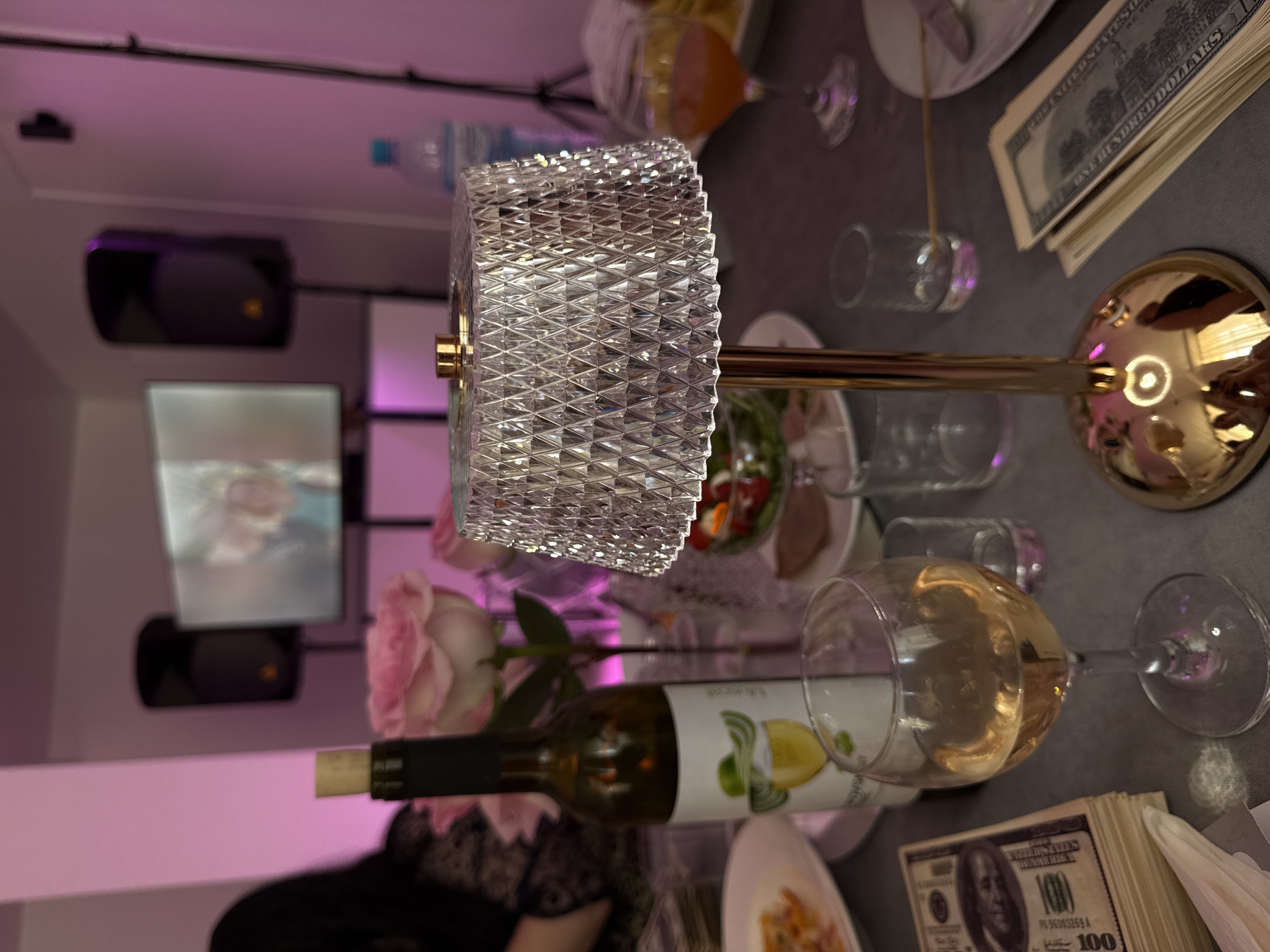 The image depicts a dimly lit indoor scene with a table setting. In the foreground, there is a stack of US dollar bills, a wine glass filled with a golden liquid, a wine bottle with a label featuring a woman in a hat and a glass of wine, and a decorative crystal lamp. In the background, there are pink roses in a vase, a television screen displaying a blurred image, and two speakers. The room is illuminated with pinkish light, possibly from ambient lighting or a light source not fully visible. The presence of the US dollar bills and the overall ambiance suggest a celebration or a gathering with a focus on luxury or a specific theme. The image does not contain any visual cues or information that would allow for the identification of the location as Moscow, Russia.