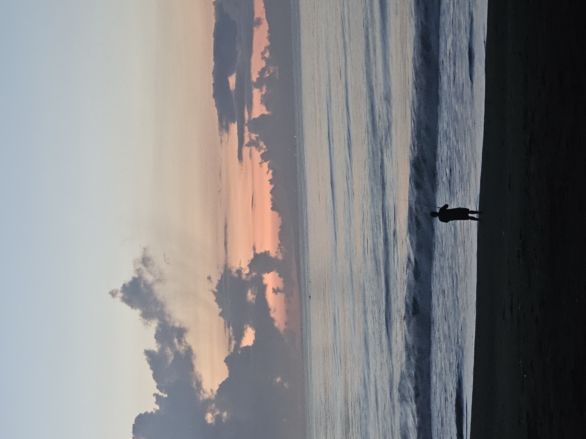 The image shows a coastal scene at sunset or sunrise, with the location likely being a beach. The sky is a mix of pale blue and orange hues, indicative of dawn or dusk, with scattered clouds. The ocean stretches to the horizon, with gentle waves rolling towards the shore. A lone silhouetted figure stands at the edge of the water, their stance suggesting they are observing the scene. The foreground appears to be sandy beach, and the overall lighting is soft, enhancing the tranquil atmosphere. The location in the city of Denpasar, Indonesia, is a likely place for this type of scene.