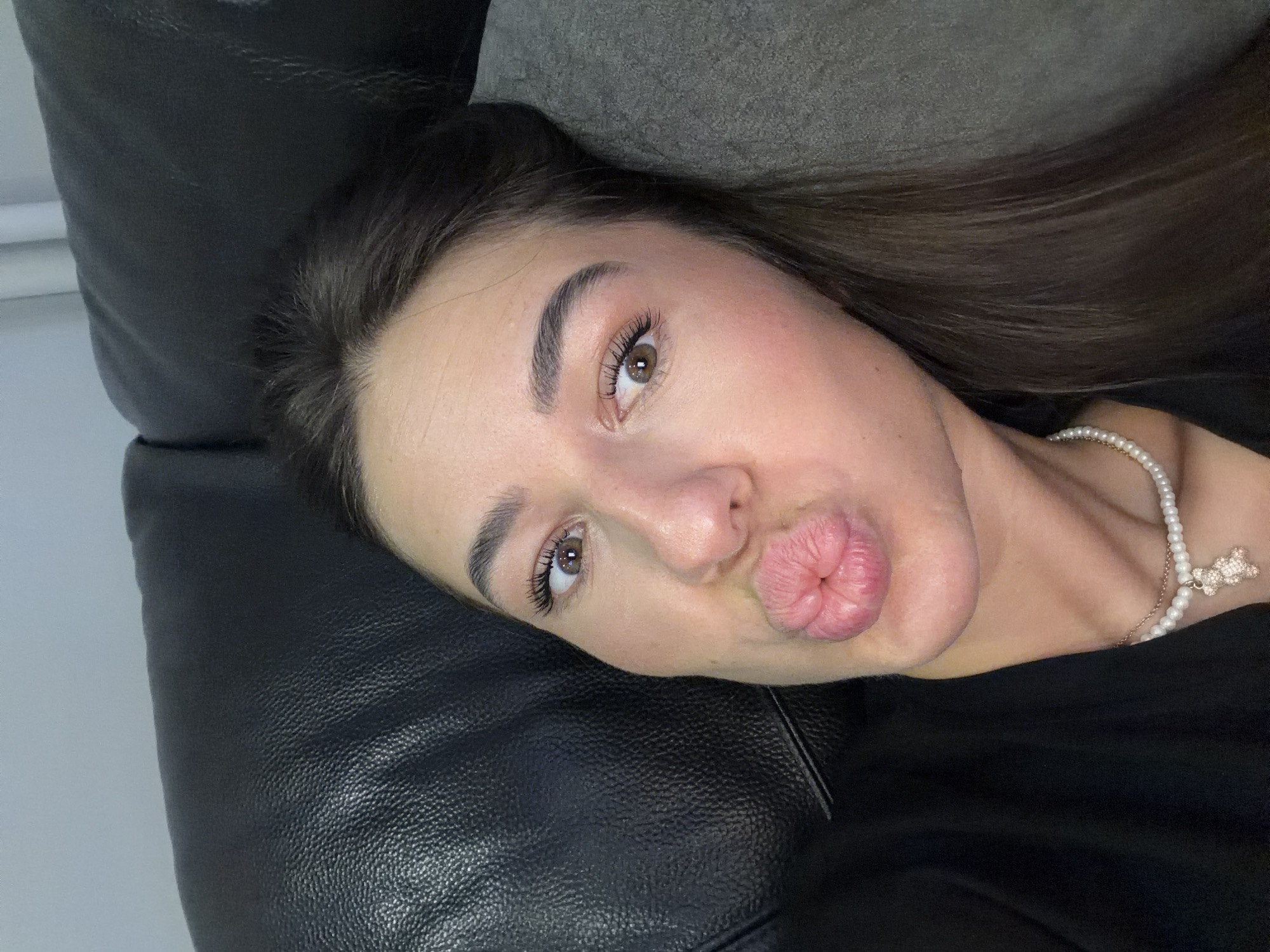 The image is a close-up selfie of a young woman with dark hair and brown eyes. She is wearing a black top and a pearl necklace with a teddy bear charm. Her lips are pursed in a kiss gesture. She is lying on a black leather couch, with a gray pillow behind her head. The background appears to be a wall. There is no indication of the city of Novi Iskar, Bulgaria, in this image.