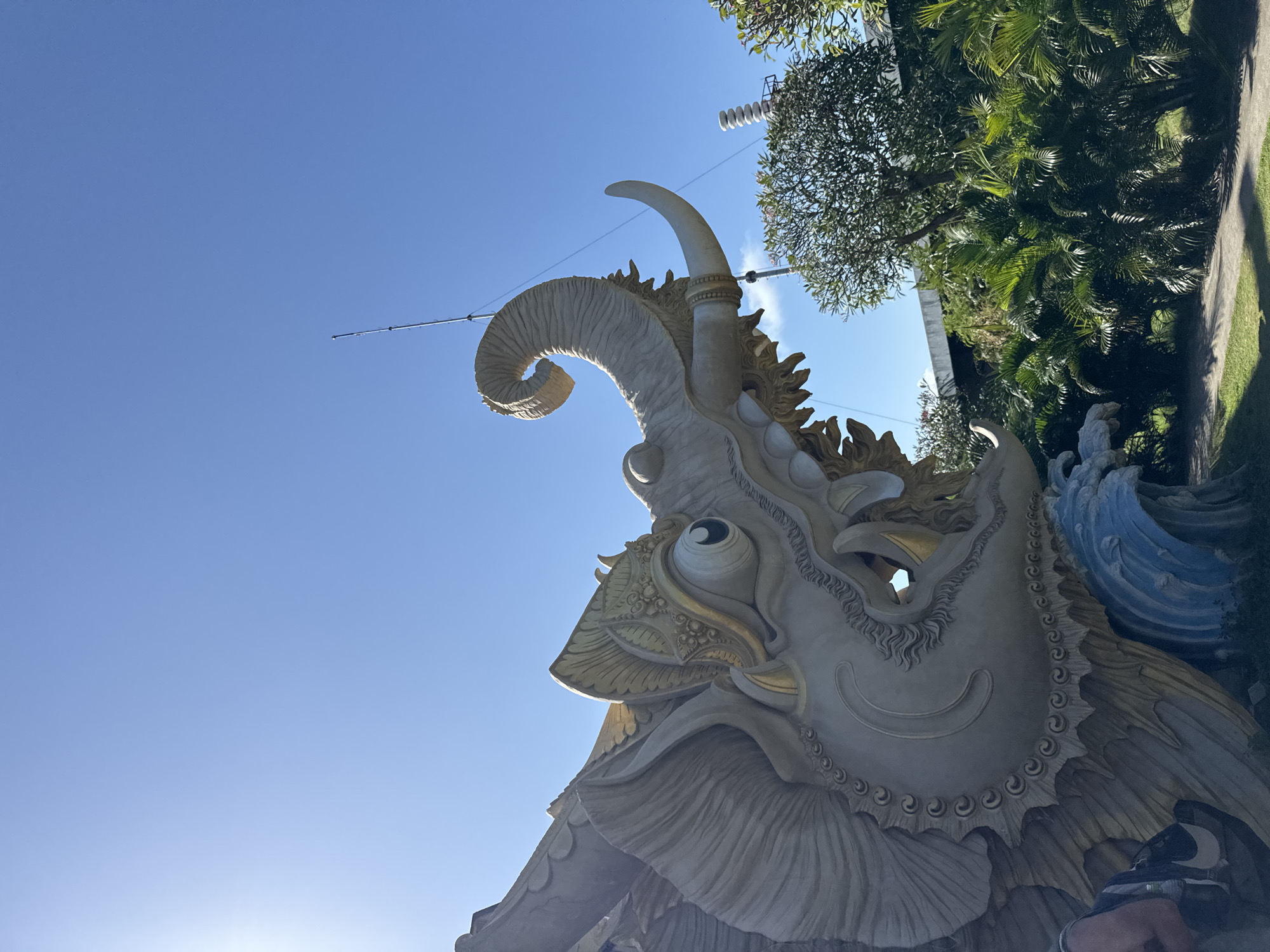 The photograph captures a close-up, low-angle view of a large, ornate statue resembling a mythical elephant with wings, set against a clear blue sky. The statue is predominantly beige with gold accents, particularly around its elaborate headdress and facial features. Its trunk is curled upwards, and it possesses a single tusk. Part of its body appears to be merging with a sculpted blue wave at the base.
In the background, lush green foliage, including palm trees, is visible, suggesting a tropical or subtropical environment. There is a hint of a modern structure, possibly industrial, with stacked elements, and a tall antenna or pole reaching into the sky. Sunlight beams in from the upper left, casting light on the statue and creating shadows. A small section of a person, wearing a backpack with a Nike logo, is visible on the far left of the frame, suggesting the statue is a tourist attraction.
This statue is identified as the "Garuda Wisnu Kencana" (GWK) statue, a prominent landmark located in the southern part of Bali, near the city of Denpasar, Indonesia. The statue is a significant cultural and tourist attraction in the region.