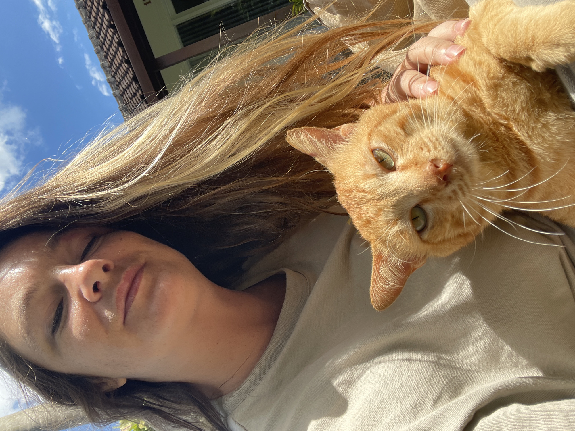 An outdoor, slightly angled medium selfie shows a woman holding an orange tabby cat. The woman's face is partially visible, with her head tilted down and eyes closed, bathed in sunlight. Her long, wavy blonde and brown hair flows around her head and shoulders. She is wearing a light-colored t-shirt. The cat, also bathed in sunlight, looks directly at the camera with green eyes and long white whiskers. Its orange fur is visible along with its ears and front paws. The background includes a tiled roof and blue sky with white clouds, suggesting a residential area. The woman's hand is resting on the cat's back. The scene appears to be in North Kuta, Indonesia.