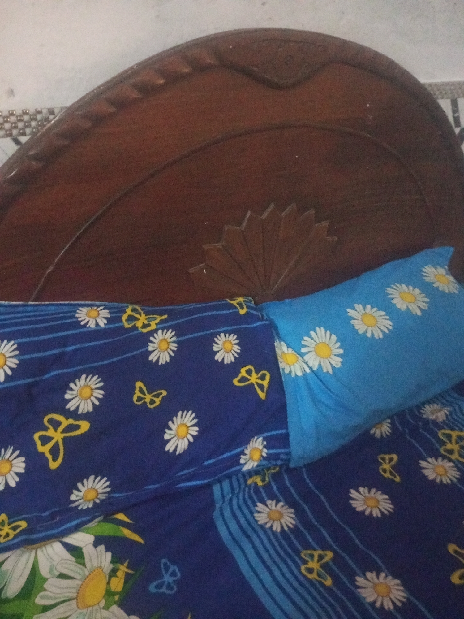 A close-up, low-angle interior shot captures a bed with a dark wooden headboard adorned with carved floral motifs and geometric patterns. A blue pillow with white daisy patterns is placed on the bed, next to a dark blue duvet cover also decorated with white daisies and yellow butterflies. The bed is positioned against a textured, off-white wall. The overall scene suggests a domestic setting, possibly a bedroom, within a home. The image is devoid of any individuals or any explicit indication of location within Mansehra, Pakistan.