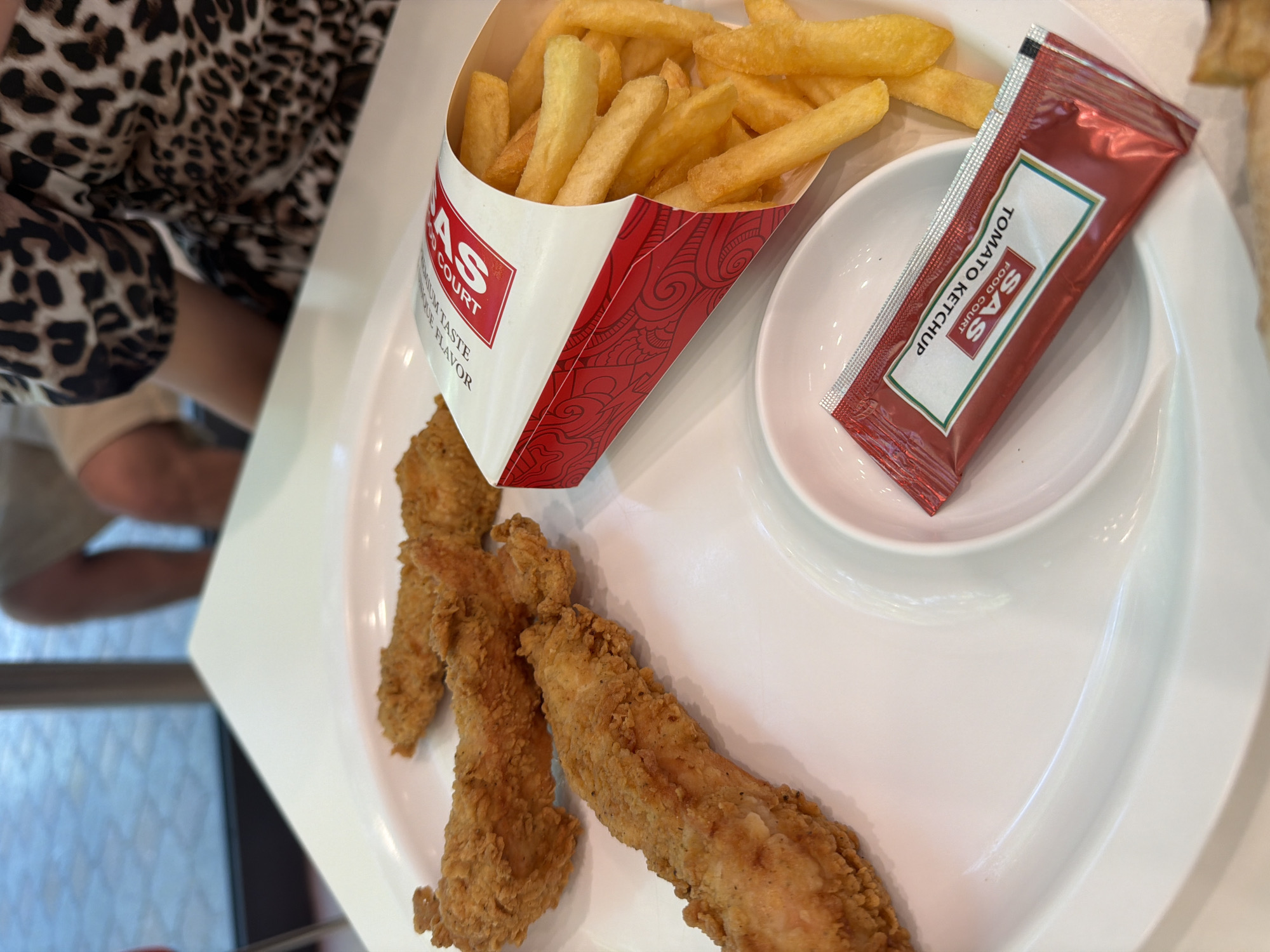 A plate of fried chicken tenders and French fries sits on a white table. The chicken tenders are golden brown and appear crispy. The French fries are also golden brown and are served in a red and white paper container. A packet of tomato ketchup is placed on the side, on a small white dish. In the background, a person wearing a leopard-print top is visible. The scene appears to be a casual dining setting, possibly a fast-food restaurant. The lighting is bright and natural, suggesting the photo was taken during the day, perhaps near a window as indicated by the blurred outdoor scenery visible through a glass pane in the background. The location in Yerevan, Armenia, is not discernible from the visual information provided in the image.