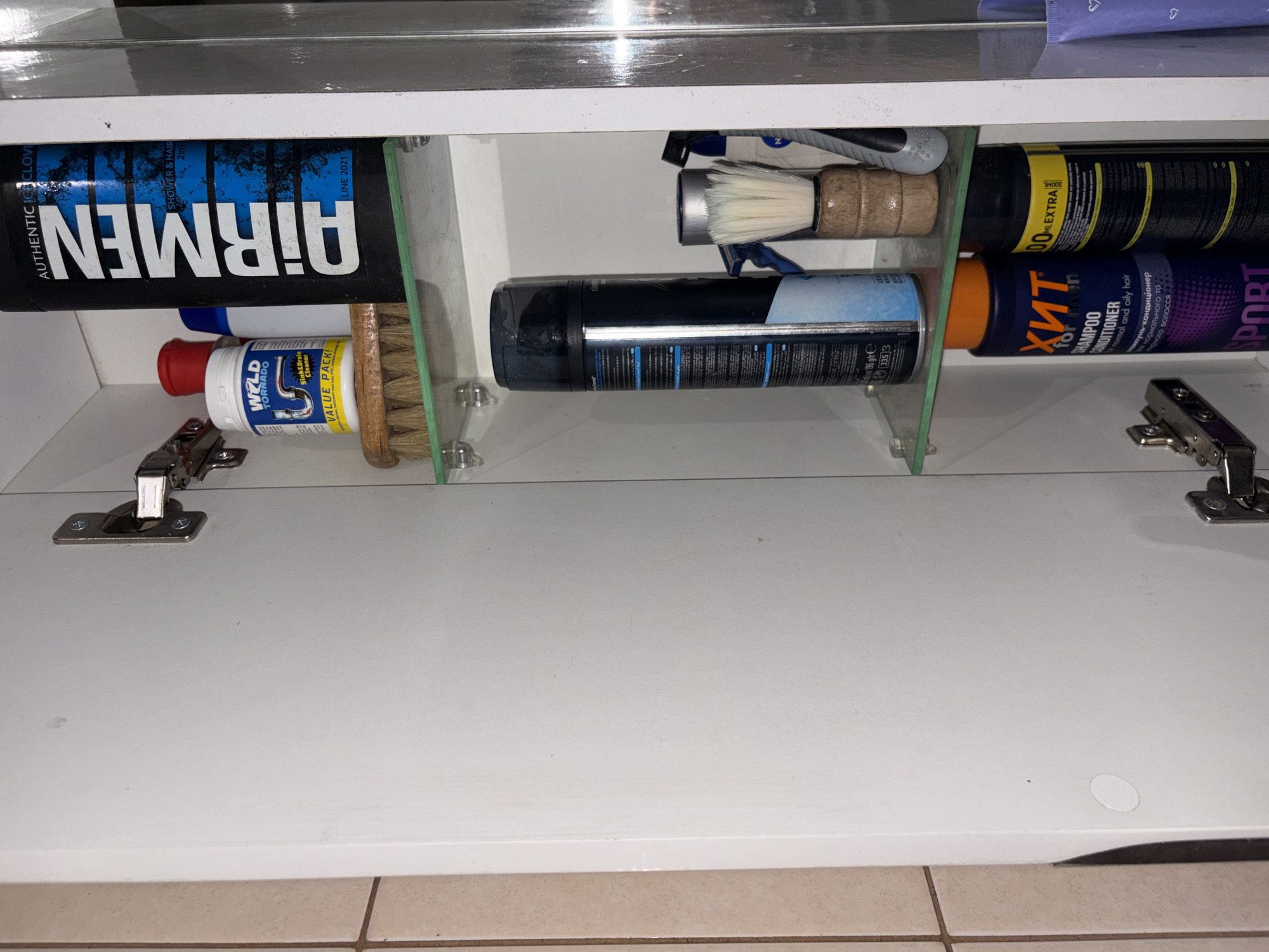The image shows the interior of a white medicine cabinet with shelves. On the top shelf, from left to right, are a white plastic bottle with a red lid, a white bottle labeled "WELD TORNADO SINK & DRAIN CLEANER VALUE PACK!", a blue and white bottle, and a tall black bottle with blue lettering that says "AIRMEN SHOWER & HAIR 2 IN 1 LINE 2021". A wooden-handled brush with bristles is positioned in front of the "WELD TORNADO" bottle.
On the second shelf, a large black can with blue and white text and graphics is standing upright. To its right is a small wooden-handled shaving brush with white bristles. Behind the shaving brush are the tops of other grooming products.
On the third shelf, two large bottles are visible. The one on the left is dark blue with an orange top and has "ХИТ for men SHAMPOO CONDITIONER" written on it. The bottle to its right is black with a yellow label that says "100 ML EXTRA".
The cabinet has a white interior and is equipped with hinges on the left side. The overall environment appears to be a bathroom or personal grooming area. There are no people or animals visible in the image.