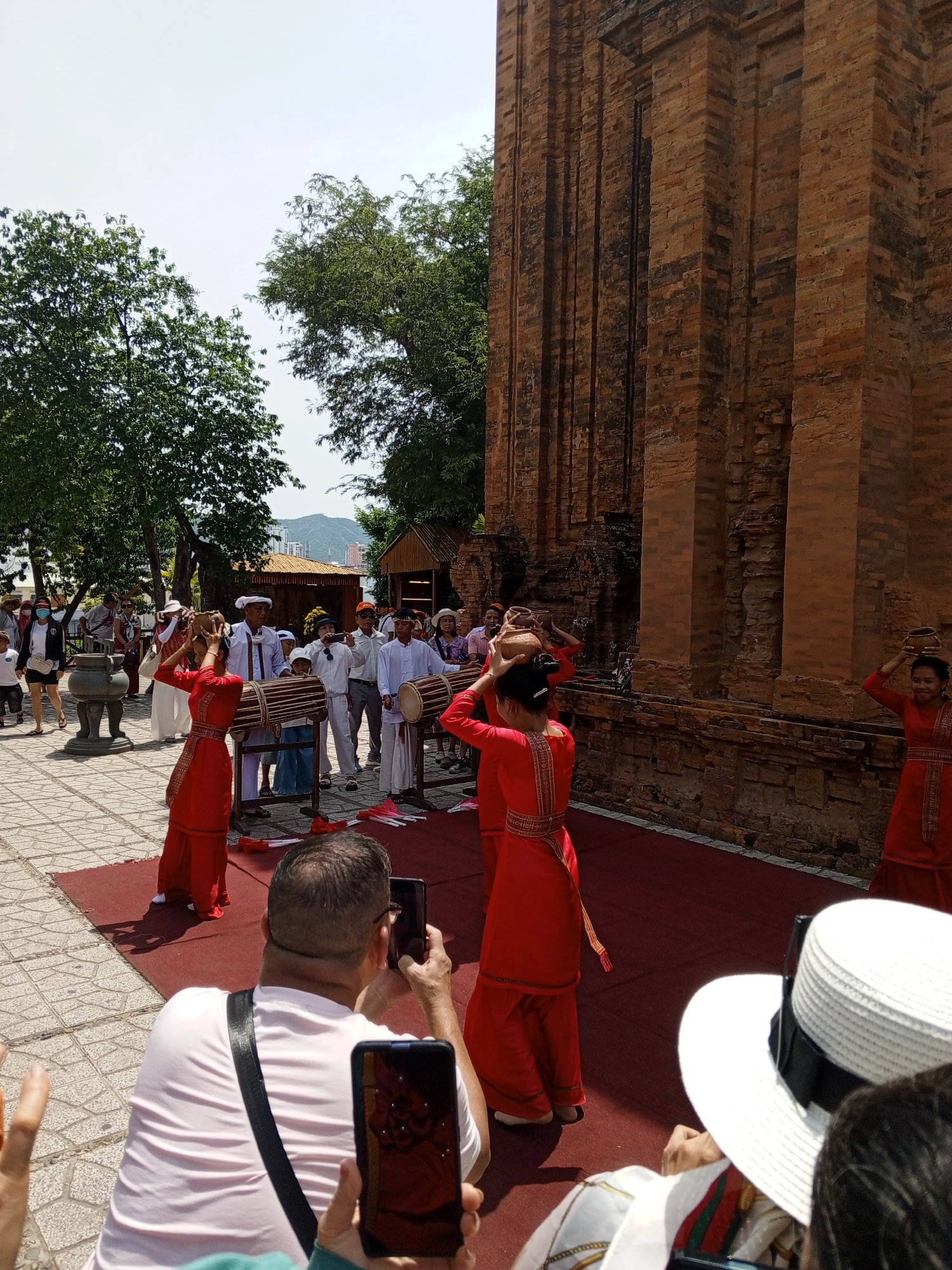 In the city of Nha Trang, Vietnam, a group of people are gathered in an outdoor courtyard. The central area is covered by a red carpet, upon which several individuals are engaged in a performance. Two women in vibrant red traditional Vietnamese dresses are performing a dance, each balancing a clay pot on their heads. They are accompanied by a group of men also in traditional attire. Some men are playing large cylindrical drums that are mounted on stands, while others appear to be watching the performance. A large brick tower, characteristic of Cham architecture, dominates the background. In the foreground, several spectators are observing the event, with some taking photographs with their mobile phones. The atmosphere appears to be festive, with clear skies and lush trees providing a backdrop to the cultural display. The location is confirmed by visual cues and architectural style, typical of Cham towers found in and around Nha Trang.
