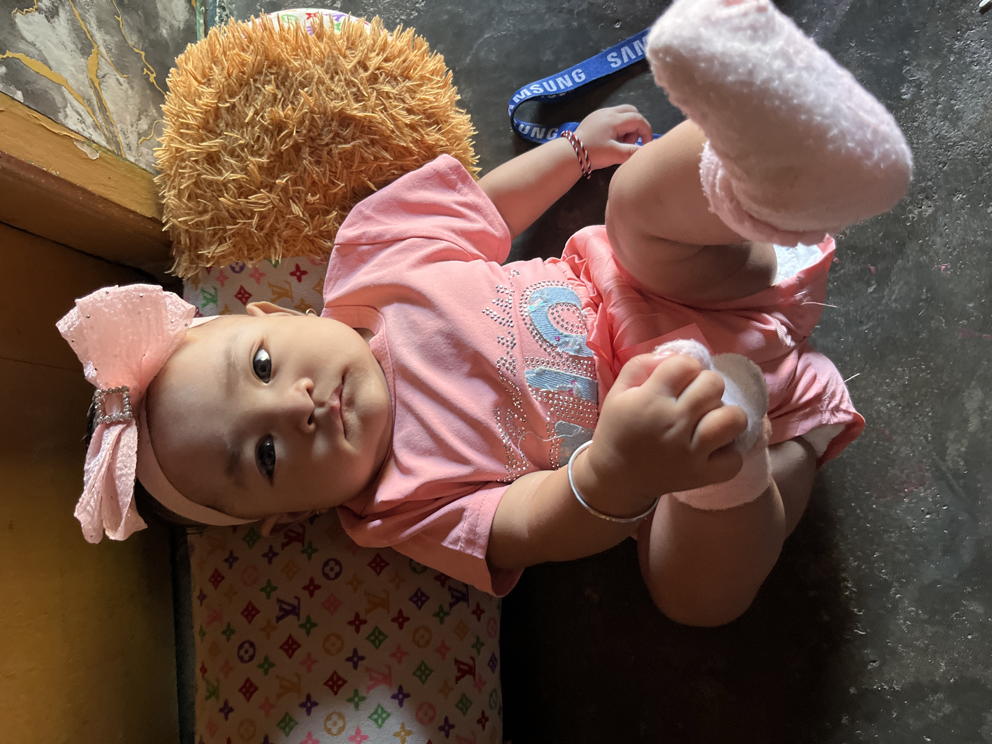 A baby girl is sitting on a dark floor in what appears to be a residential setting in Kruengmate, Indonesia. She is wearing a pink outfit consisting of a t-shirt and shorts, adorned with sparkly embellishments on the shirt. A large pink bow with a jeweled accent is worn on her head. The baby has a silver bracelet on her left wrist and a red and white braided bracelet on her right wrist. She is holding a white fabric object in her hands. To her left is a patterned pillow with a repeating logo, and to her right is a shaggy brown cushion. A blue strap with white lettering, partially visible, is present in the background. The environment appears to be indoors with a yellow wall and wooden elements visible in the upper portion of the frame.