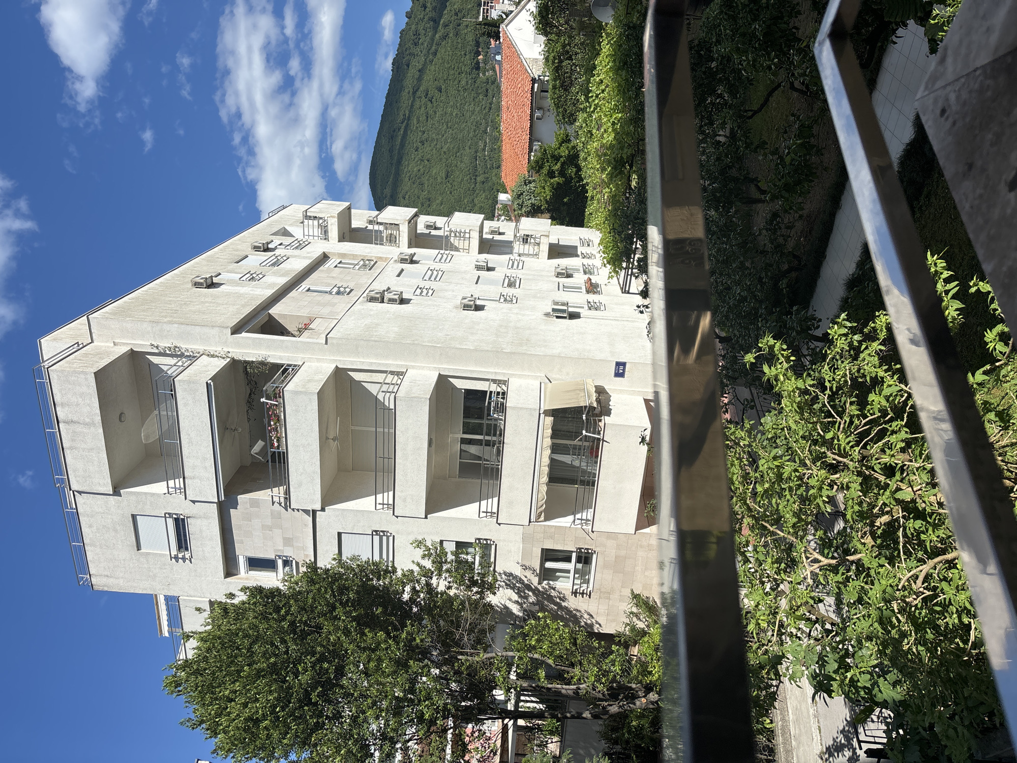 The image shows a modern, white apartment building in Budva, Montenegro, viewed from a slightly elevated position behind a metal railing. The building has several floors with recessed balconies and numerous air conditioning units. A tree partially obscures the lower floors. In the background, a mountain rises, and a building with a red tile roof is visible. The sky is blue with scattered white clouds. The photograph is slightly rotated.