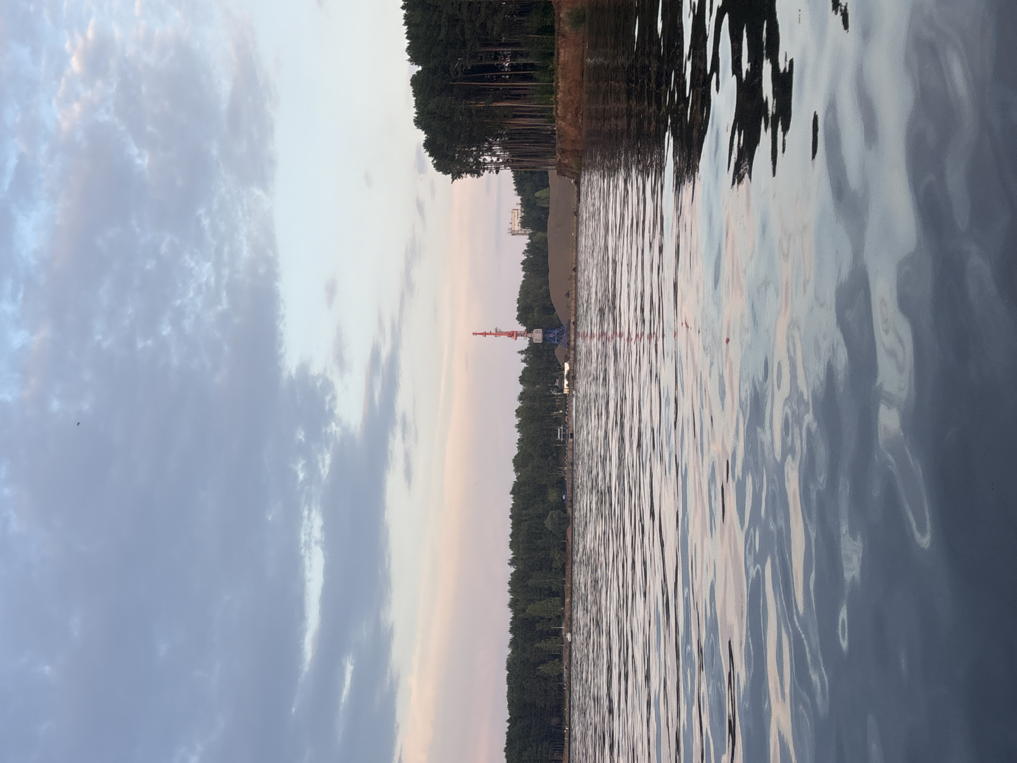 This image depicts a wide body of water, likely a river or reservoir, with a shoreline lined by a dense forest of tall, dark green trees. The water's surface is gently rippled, reflecting the soft, pastel colors of the sky, which appears to be either sunrise or sunset, with hues of pink, blue, and gray.
In the mid-ground, on the left side of the frame, a large industrial crane with a red boom stands prominently against a backdrop of sand or earth mounds. Beyond the mounds and trees, a tall, light-colored building is visible in the distance. On the right side of the image, the shoreline is characterized by a steep, reddish-brown embankment with pine trees growing along its edge.
The scene is tranquil and natural, with no visible people. The presence of the crane and the sand mounds suggest some form of industrial or construction activity near the water. The location is likely on the Kama River in Chaikovsky, Russia, given the characteristic industrial elements juxtaposed with the natural landscape. The overall atmosphere is serene, bathed in the diffused light of dawn or dusk.