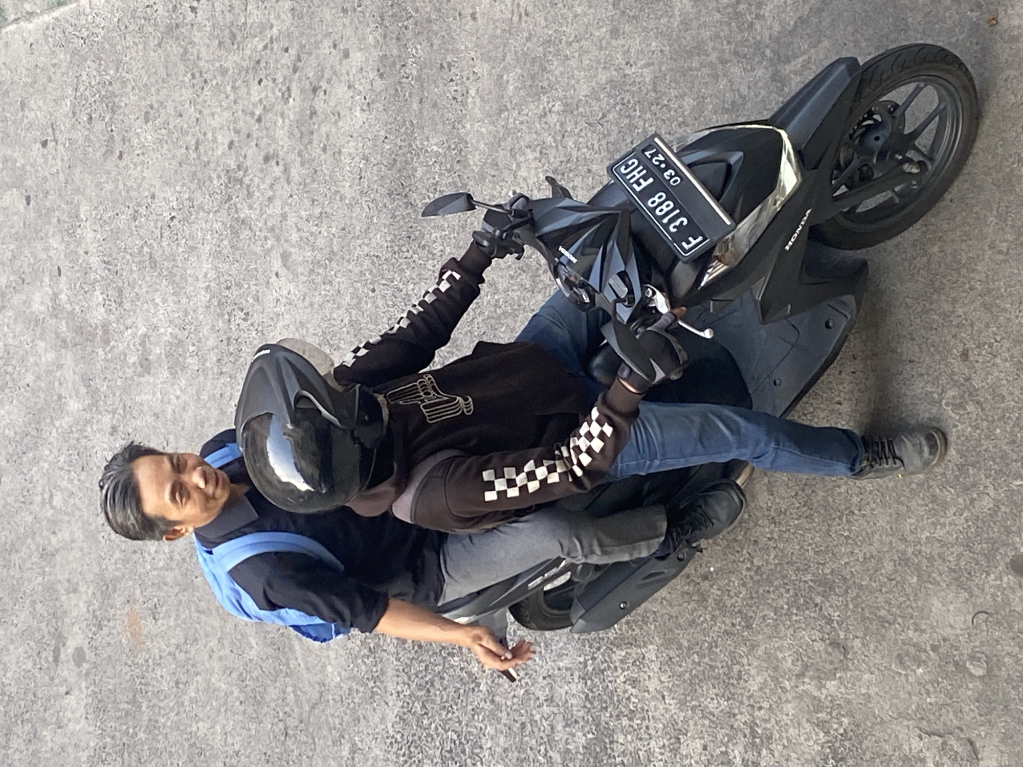 Two individuals are riding a black Honda scooter on a paved road, likely in North Jakarta, Indonesia, given the license plate "F 3188 FHG 03-27". The person in front, wearing a black helmet and a black hoodie with white checkered sleeves, is operating the scooter. The person in the back, wearing a black shirt and a blue backpack, is seated behind the driver and is holding a lit cigarette in their right hand. The road surface appears to be concrete with some wear and tear. The lighting suggests it is daytime.