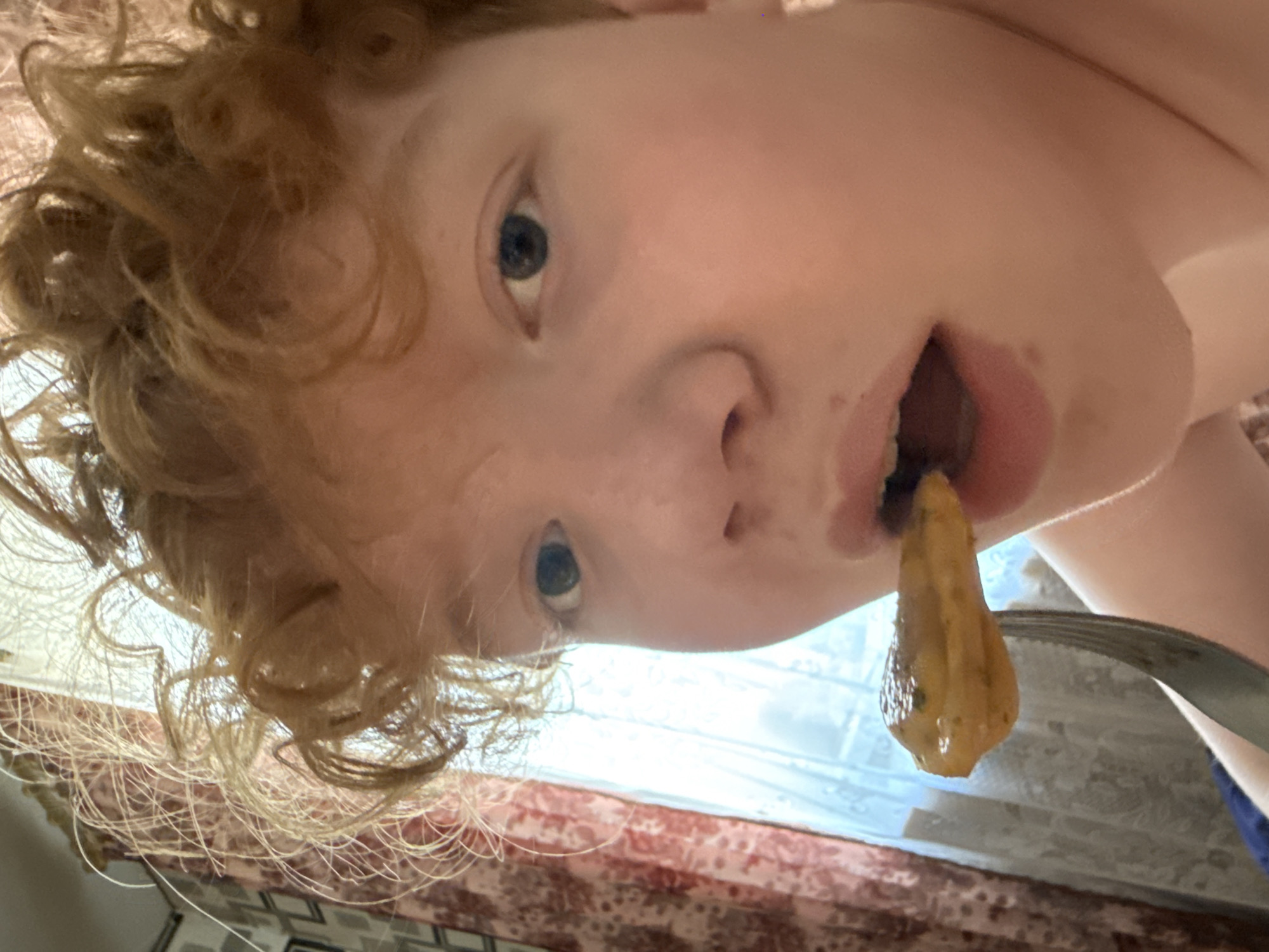 A low-angle, tilted close-up photo shows a person with light skin, blue eyes, and curly red hair holding a piece of food on a fork near their open mouth. The food appears to be yellowish-brown and somewhat textured. The background is blurry, showing what appears to be a window with a patterned curtain or blind. The location is unknown.