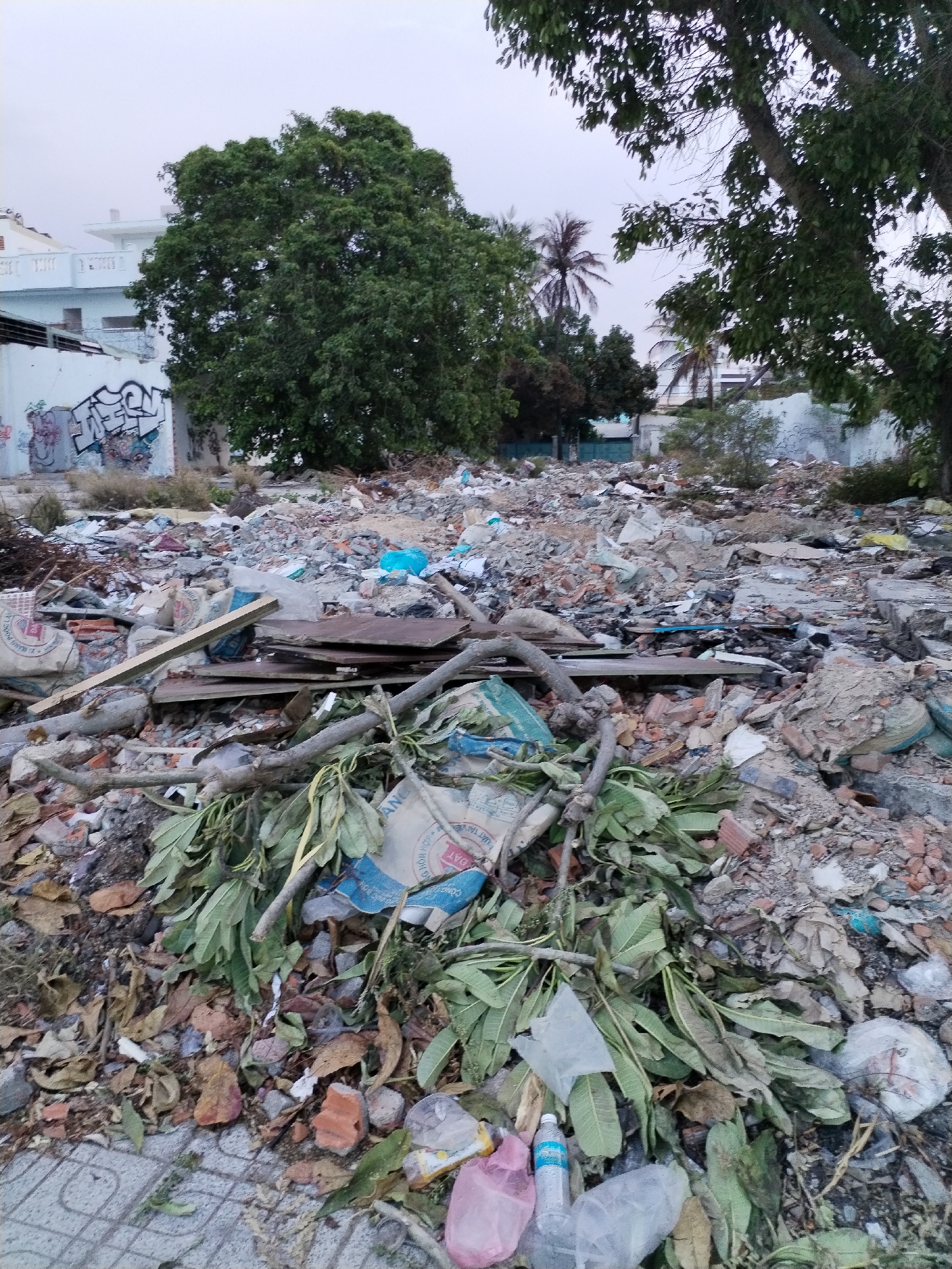 The image depicts a trash-strewn area in Bac Nha Trang Ward, Vietnam. The scene is dominated by piles of debris, including rubble, bricks, plastic bags, sacks, wooden planks, and green leaves. A concrete block wall covered with graffiti is visible in the background, with a building behind it. There are trees scattered around the trash site. The pavement can be seen in the foreground. No people or animals are visible.