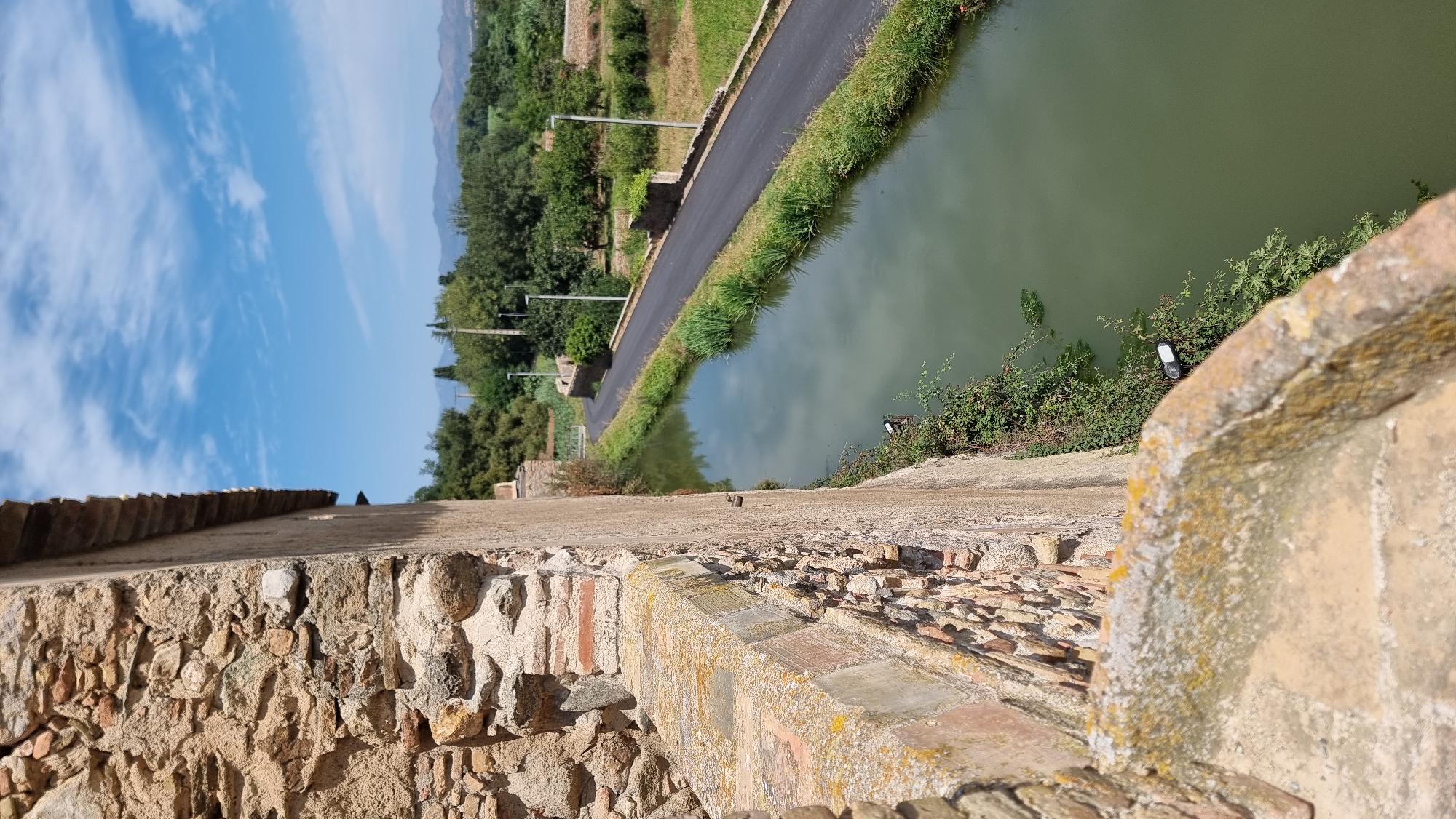 A close-up perspective, looking down from a stone wall, reveals a canal on the right side of the frame, with a paved road running alongside it. The canal's water is a murky green, and lush green vegetation lines its banks. Beyond the canal and road, a verdant landscape unfolds, featuring fields, trees, and distant mountains under a bright blue sky dotted with white clouds. In the foreground, the rough-hewn texture of the stone wall is prominent, with moss and lichen clinging to its surface. A dark object, possibly a mobile phone, rests on the vegetation near the canal. The scene appears to be set in a rural area of Castelló d'Empúries, Spain.