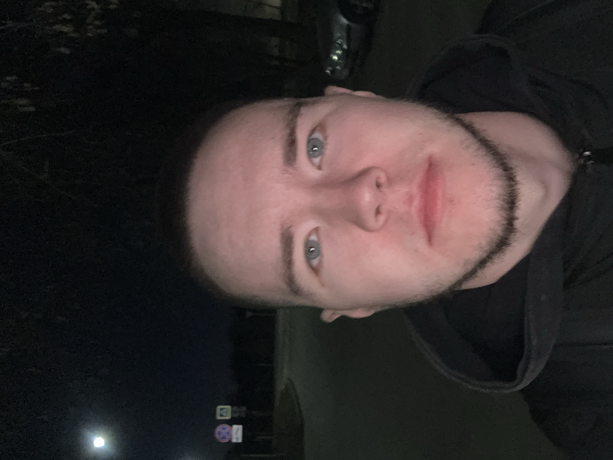 A close-up, vertical portrait captures a young male individual outdoors at night in Kazan, Russia. The subject has fair skin, light blue-grey eyes, dark eyebrows, and a chin strap beard. He is looking directly into the camera with a neutral expression, showing a slight smirk. He wears a dark, hooded garment.
The setting is dimly lit, indicating nighttime. In the background, various dark elements are visible, including what appears to be foliage or buildings and a partial view of a vehicle at the top right. Towards the bottom left, there's a bright, artificial light source, possibly a street lamp or vehicle headlight. Below the subject's left eye and slightly to the left, two road signs are visible on a pole. The top sign is a blue square with a white pedestrian symbol, common for a pedestrian crossing. The bottom sign is round, purple/pink with white elements, but too blurry to decipher its specific meaning. The overall atmosphere is dark, with the subject's face being the primary illuminated area.