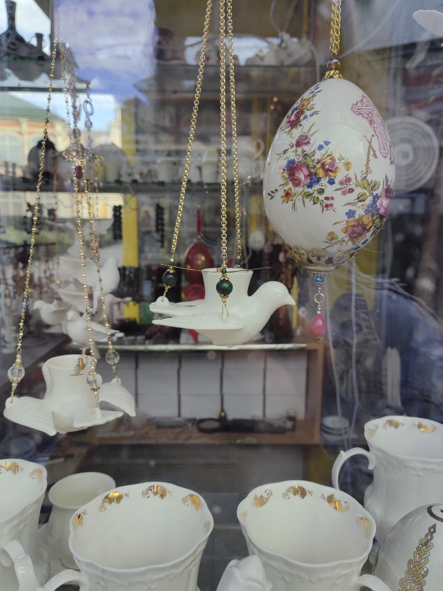 This image captures a close-up view of a display of decorative porcelain items, likely within a shop window in Saint Petersburg, Russia. The primary subjects are several white porcelain pieces, including what appear to be cups or pitchers adorned with delicate gold detailing, and several decorative bird figurines.
In the center of the frame, a white porcelain bird is suspended by a gold chain, holding a small white dish. Above it, a large, ornate white egg-shaped ornament hangs from another gold chain, decorated with a vibrant floral pattern in shades of pink, blue, and yellow, and what looks like a pink cross and the letter "N" with gold accents. To the left and right, more delicate gold chains with small beads and charms are visible, hanging alongside other white porcelain items, including more bird figures. The reflections in the glass window suggest an outdoor setting, possibly a street scene with buildings and possibly clouds visible in the sky.
The overall impression is one of fine craftsmanship and elegant decoration. The lighting suggests it could be daytime, with natural light filtering through the window. The scene appears to be static, a display intended to attract customers. There is no visible text within the frame that is legible. The focus is entirely on the intricate details of the porcelain objects.