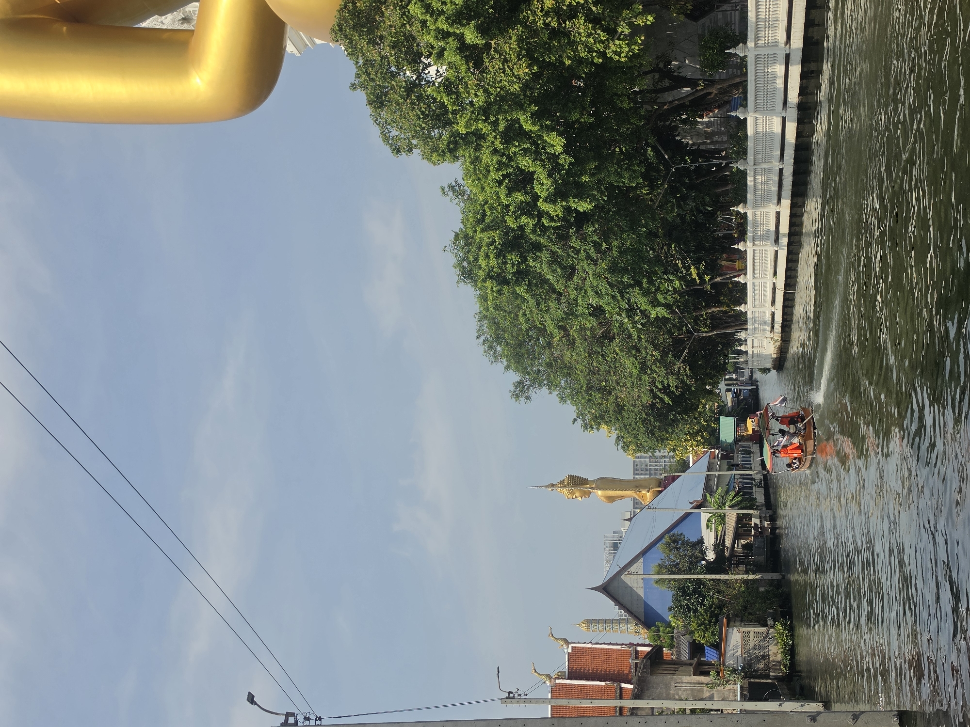 The image shows a canal in Bangkok, Thailand. A long-tail boat with several people wearing orange life vests is traveling on the canal. Buildings and trees line the canal banks. A large reclining golden Buddha statue is visible in the background. A white decorative wall or fence runs along the water's edge on one side. Power lines stretch across the sky in the foreground.