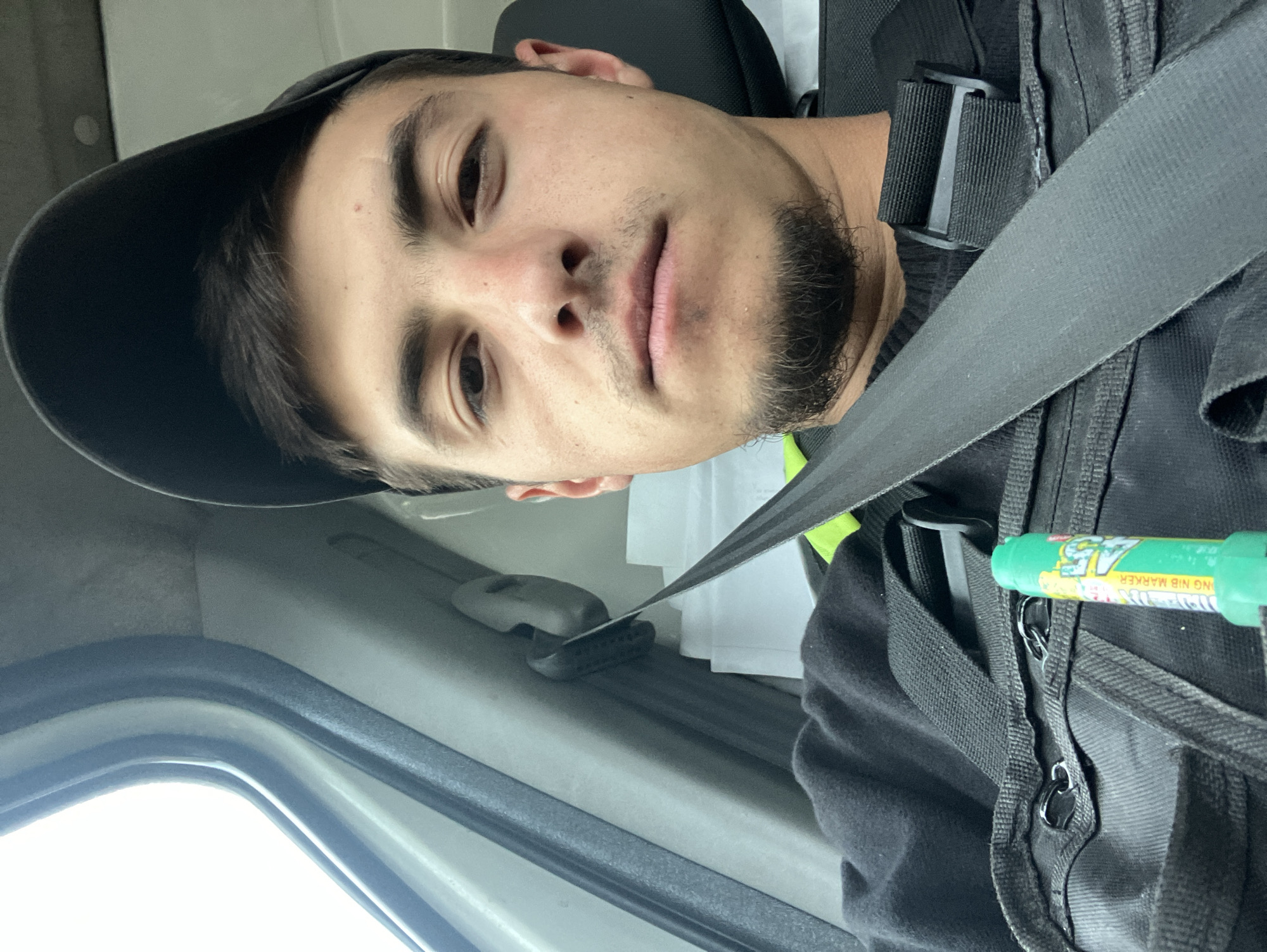 An image, rotated 90 degrees counter-clockwise, depicts a young man taking a selfie inside what appears to be the interior of a vehicle. The man, with short dark hair, a stubble beard, and dark eyes, is wearing a dark baseball cap and a dark sweater or jacket. A yellow-green safety vest is visible on his right shoulder, and he has a dark, tactical-style bag or vest strapped over his chest, with a seatbelt running across it.
His expression is neutral to slightly pensive. Behind his head, a car seat headrest is visible, along with some white papers. Through the vehicle's window on the left, a bright sky is visible, indicating it is daytime. A green marker, bearing the visible text "CHISEL NIB MARKER," is clipped to the front of his bag/vest. The interior of the vehicle shows generic grey-beige plastic trim. The man is secured by a seatbelt, suggesting he is either driving, a passenger, or the vehicle is stationary. There are no specific visual cues to confirm the location as Novo Selo, Bulgaria.