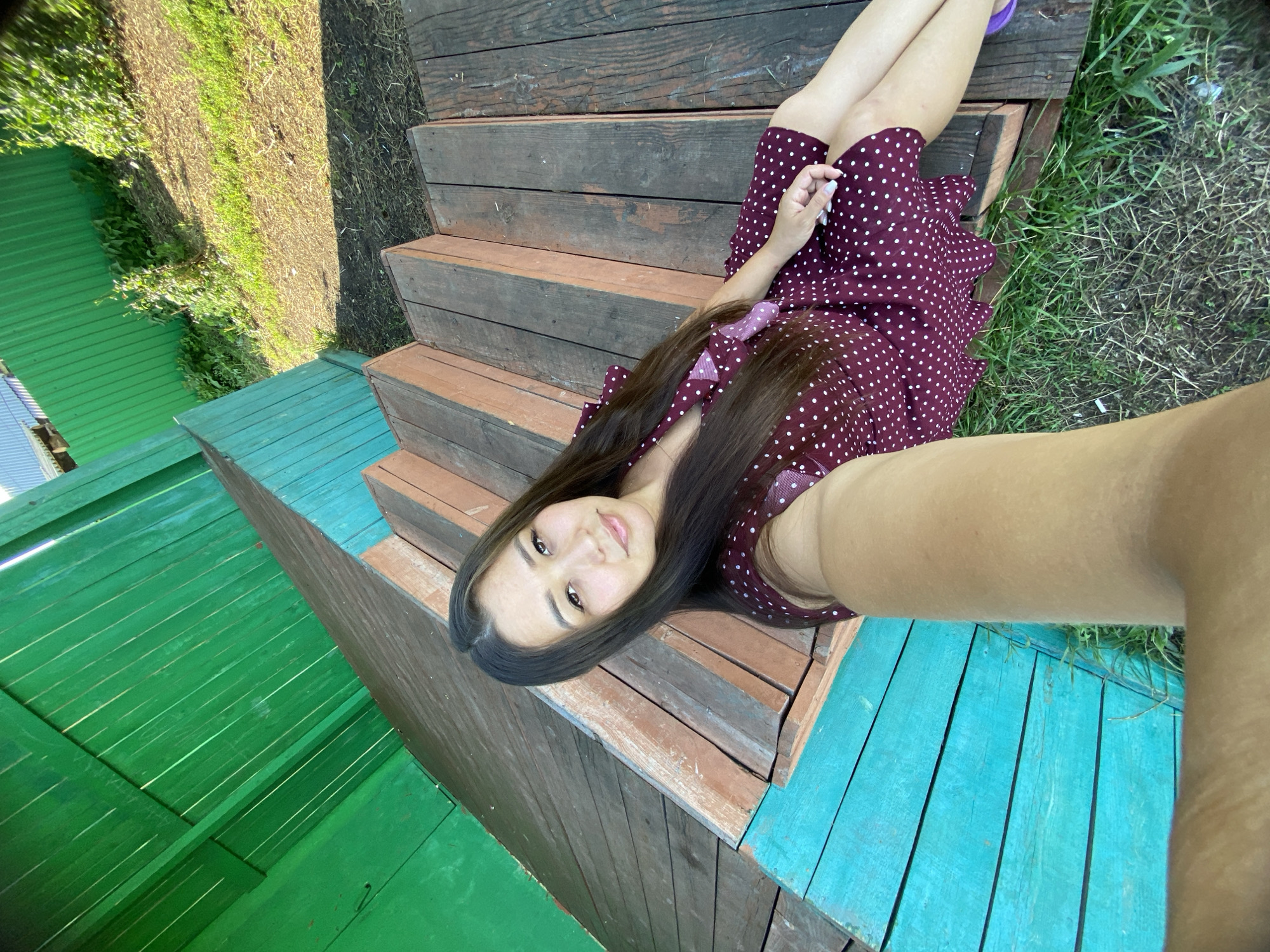 In Maloarslangulovo, Russia, a woman is captured in a selfie taken outdoors. She is sitting on wooden steps painted in teal and brown, with a green wooden fence in the background. The woman has long, dark hair and is wearing a burgundy polka dot dress. Her arm is extended, holding the phone to take the picture. The ground around the steps is covered in grass and dirt, with some foliage visible against the fence. The lighting suggests it is a sunny day.