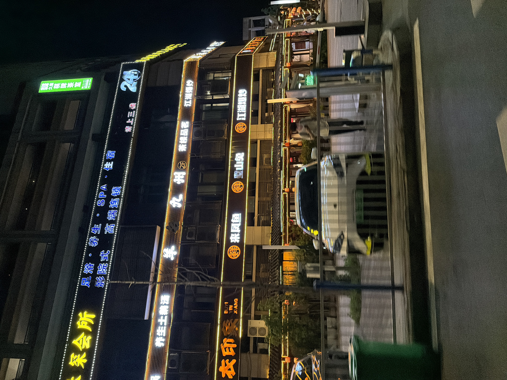 The image depicts a street scene in the Chongqing High-tech Zone, China, at night. A multi-story building dominates the scene, adorned with vertical neon signs written in Chinese characters. The signs advertise various businesses including a tea house, SPA, cinema, restaurants, and accommodation. A car and traffic barriers can be seen on the right side of the image, next to the building. The overall atmosphere is illuminated by artificial lighting from the signs.
