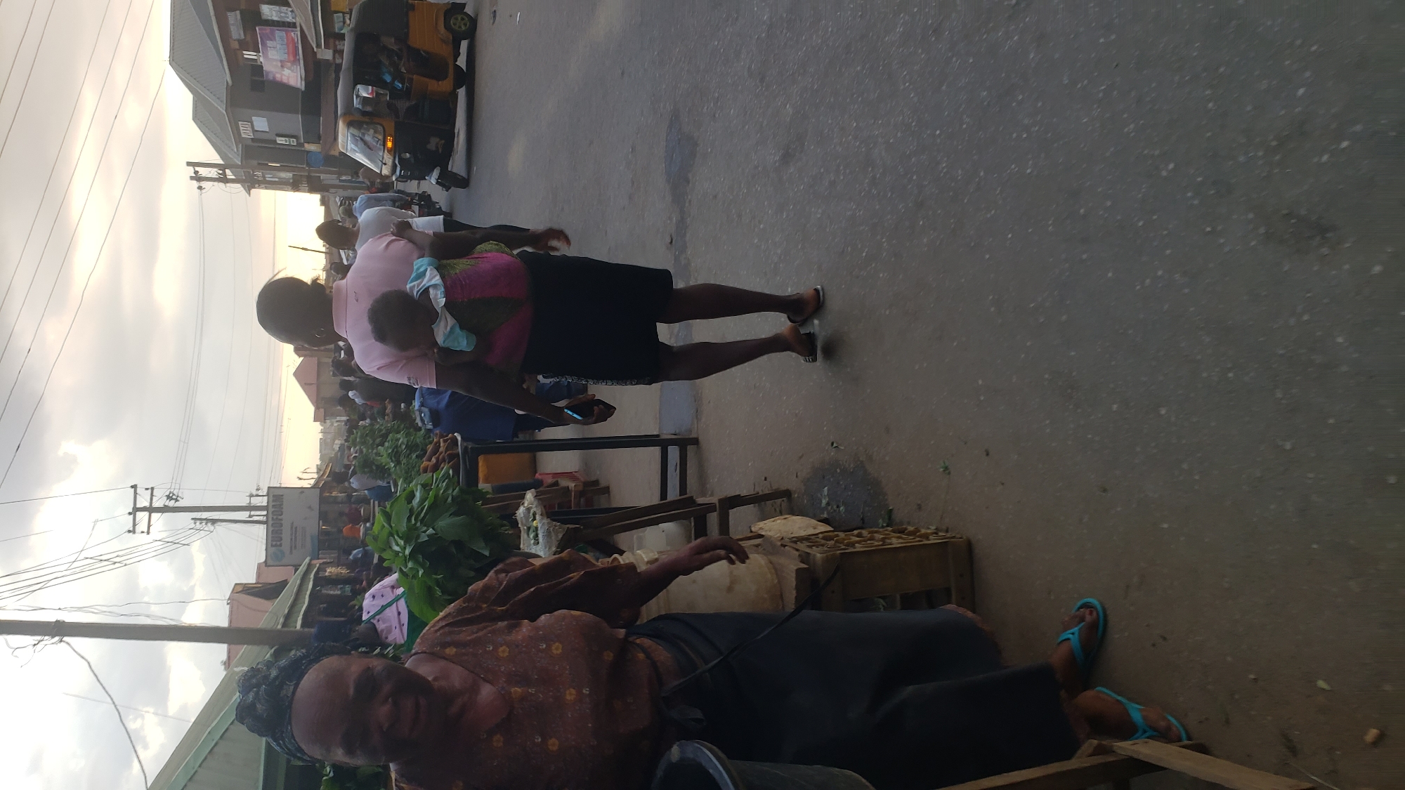 The image depicts a street scene in Narayi, Nigeria. A woman with a patterned brown top and a black skirt is seated by the roadside, positioned next to a vendor's setup of what seems to be fruit or vegetables. She is near a metal or plastic basin.
In the background, a person in a pink shirt holds a child. The street appears to be a commercial area with stalls. The background also features an autorickshaw or tricycle and overhead power lines. The street surface is asphalt.