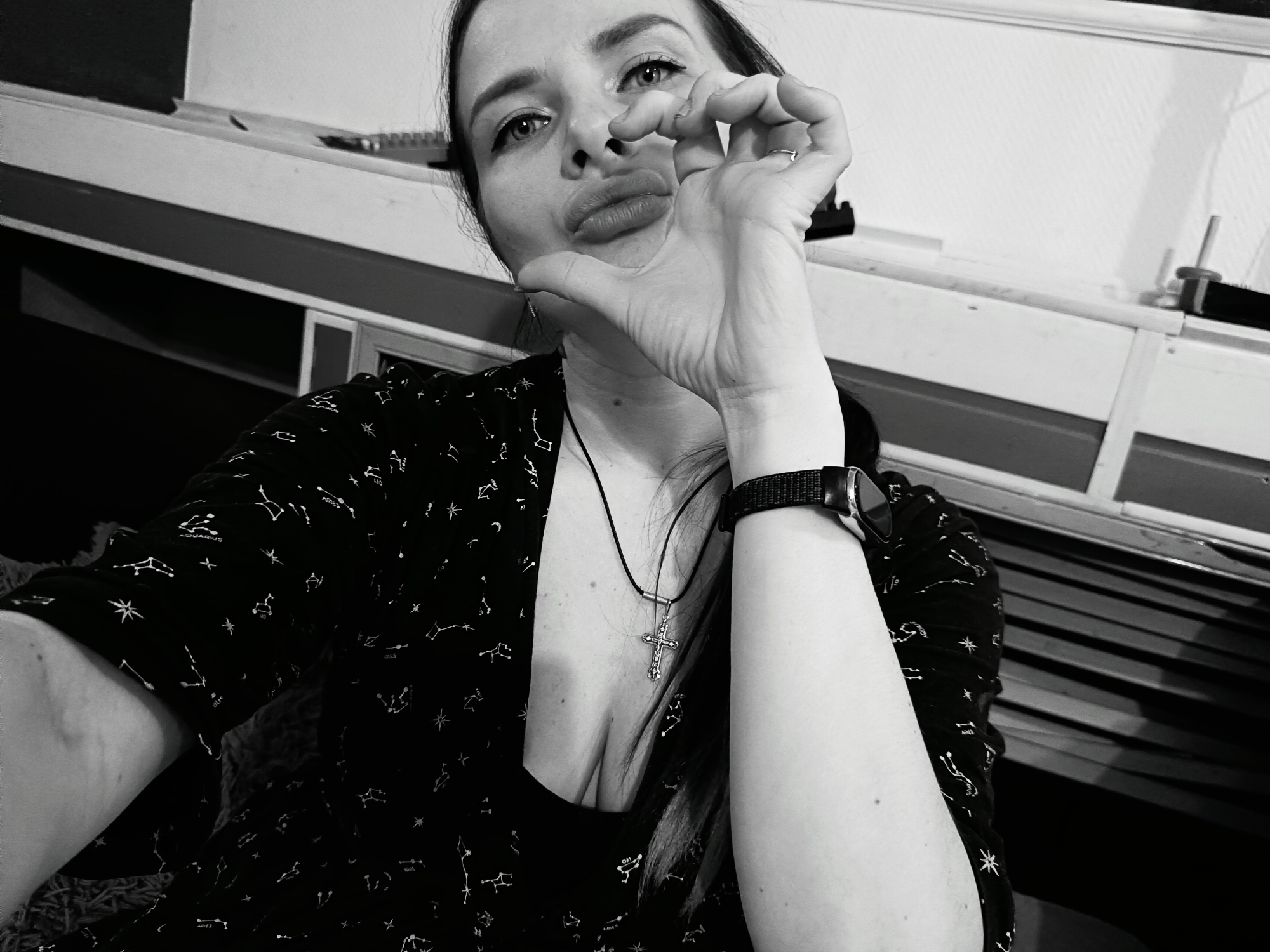 A black and white, low-angle selfie shows a woman with dark hair, looking directly into the camera with wide eyes and pouting lips. Her left hand is raised towards her face, with the index finger and thumb held close together in a pinching gesture, and the other fingers curled. She is wearing a black shirt adorned with white celestial patterns, including stars and constellation names like "AQUARIUS", "LEO", "PISCES", and "GEMINI". A black cord necklace with a silver-colored cross pendant hangs around her neck, visible in her cleavage. On her left wrist, she wears a watch with a dark strap. The background is simple, featuring a light-colored wall or shelf structure with darker horizontal elements, suggesting an indoor setting. The image conveys a playful or perhaps sassy mood through her expression and pose. The scene is located in Rusalevka, Russia.