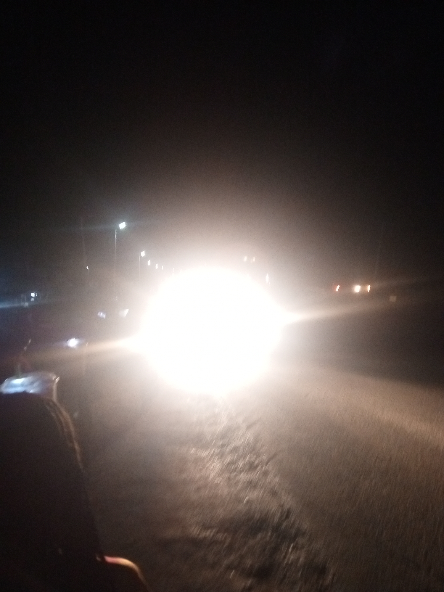 The image shows a dark, nighttime scene, likely on a road, with strong headlights illuminating the foreground and obscuring details. A person's head and shoulder are visible on the left side of the frame, appearing to be looking forward. The road surface is visible and appears wet or reflective, with streaks of light suggesting movement. In the background, streetlights and other vehicle lights are visible, creating a blurred and hazy atmosphere. The overall impression is one of motion and low visibility, common in night driving conditions. The location is unspecified.