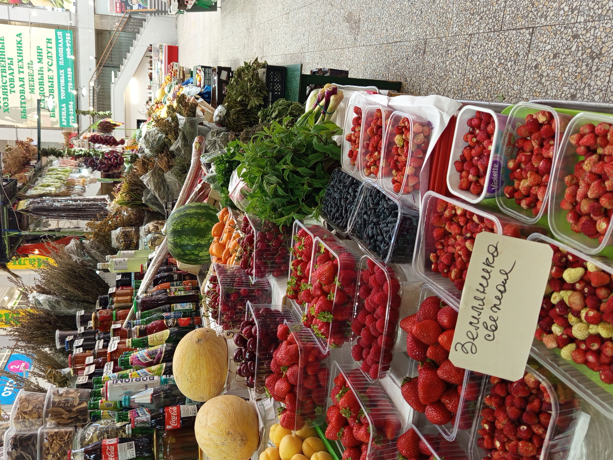 This is an eye-level, slightly tilted shot of a vibrant outdoor market stall. The foreground is dominated by an array of fresh produce, including large, ripe strawberries, raspberries, cherries, and two melons, displayed in clear plastic containers. Behind the fruit, various dried herbs and plants are bundled together, alongside bottles of liquids and jars of preserved goods. The backdrop reveals more market stalls with a variety of items, including what appears to be bottled beverages and dried foods. A prominent green sign with white Cyrillic text is visible on the left, listing types of goods and services. The floor of the market appears to be paved with grey tiles. The overall impression is a bustling, colorful marketplace offering a wide selection of fresh and preserved goods. The text "Coca-Cola" is visible on several bottles. A handwritten sign in Cyrillic, translating to "Wild strawberries, fresh," is placed in front of a container of strawberries. The lighting suggests it is daytime, and the setting is consistent with a market in Saint Petersburg, Russia. No people are clearly visible in the image.
