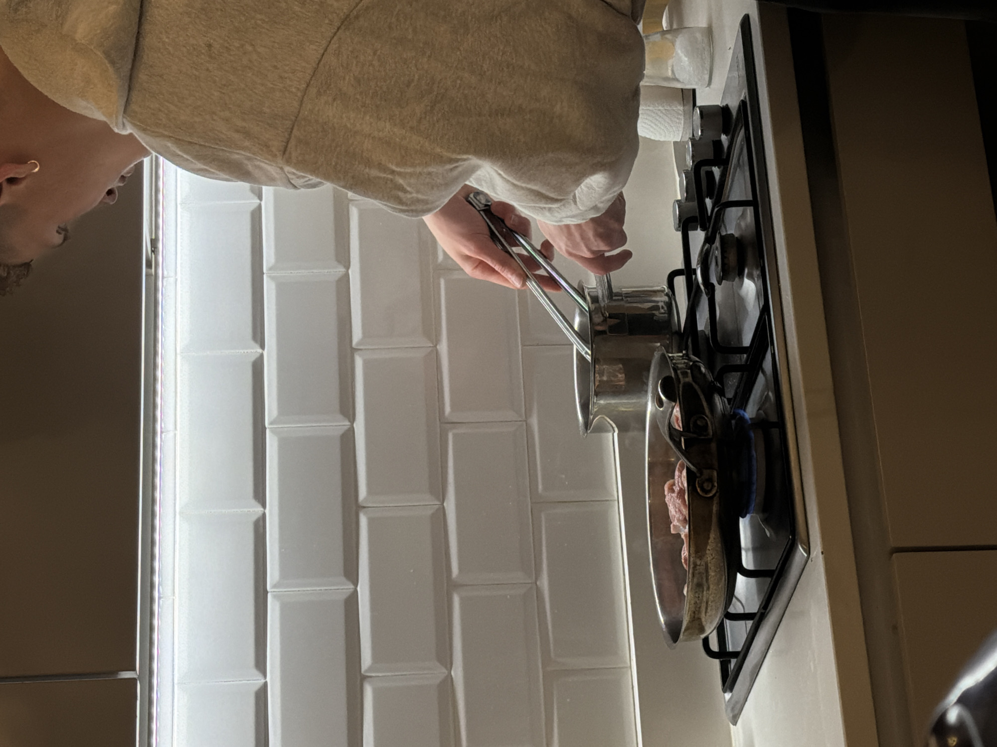 A person wearing a grey hooded sweatshirt is using metal tongs to cook meat in a stainless steel pan on a gas stove. The stove is white with black grates and is located in a kitchen with white subway tile. The location is likely London, United Kingdom.