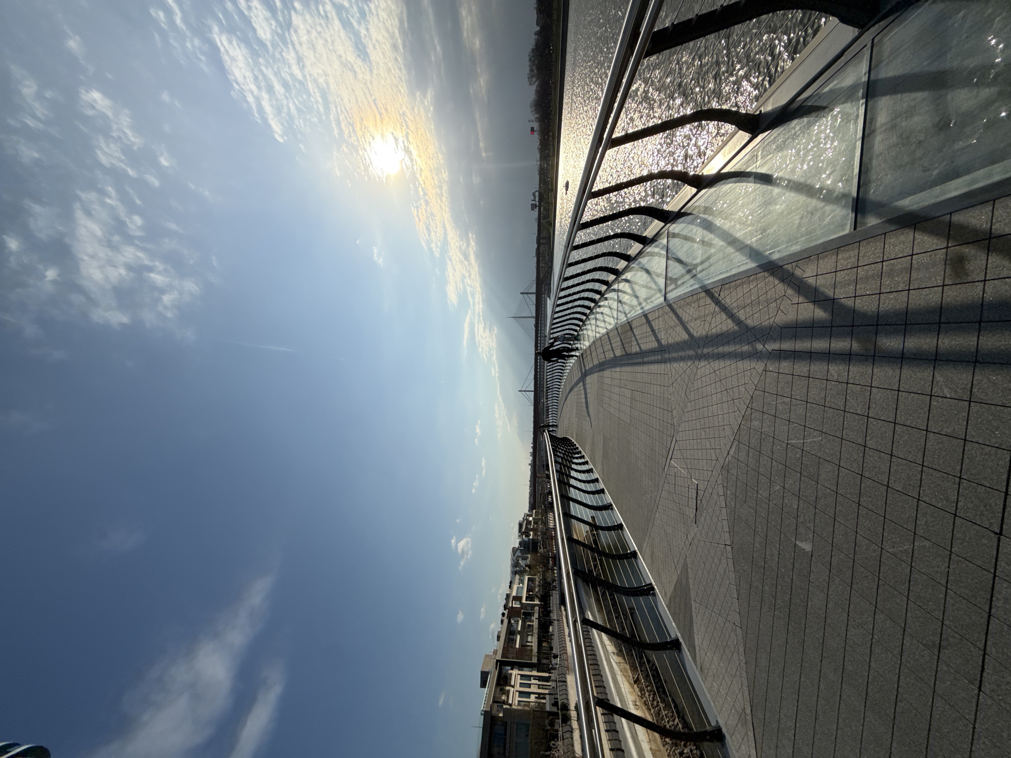 The image depicts a curved pedestrian promenade along the Sava River in Belgrade, Serbia, specifically within the Belgrade Waterfront development. The promenade is surfaced with grey rectangular paving tiles arranged in alternating patterns. A modern railing, composed of multiple horizontal stainless steel bars supported by dark, curved vertical posts, lines both edges of the walkway. On the right side, between the railing and the river, a segment features clear glass panels.
The Sava River occupies the right half of the scene, its surface rippled and brightly reflecting the sun. A small, white, indistinct vessel or object is visible on the river in the mid-distance. Along the left bank, a series of multi-story, contemporary buildings, characteristic of the Belgrade Waterfront, are visible. In the far distance, the distinctive cable-stayed Ada Bridge spans the river, with its pylon and cables prominent against the skyline. Another bridge, possibly a railway bridge, is also discernable further away.
The sun is positioned in the upper right quadrant of the sky, partially obscured by scattered white and grey clouds, casting a strong glare and emitting visible sun rays. This illumination creates long, distinct shadows of the railing and a walking figure across the promenade surface, extending from left to right. One individual is visible in the mid-ground, walking away from the camera on the promenade, appearing as a dark silhouette due to the backlighting. The sky above is predominantly blue with varying cloud formations.