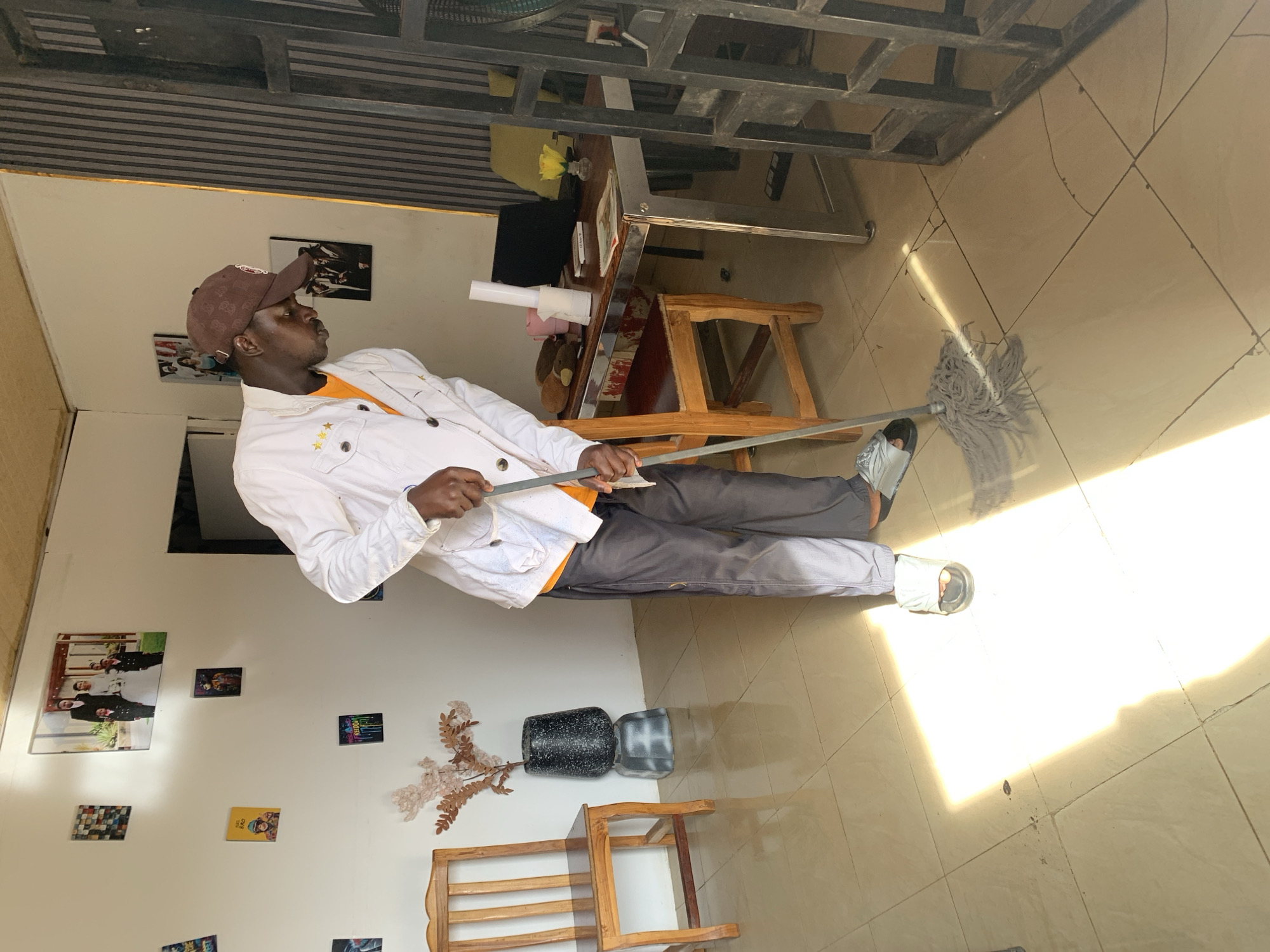 A man is depicted mopping a tiled floor in an indoor setting, illuminated by strong natural light. He stands centrally, holding a long-handled mop with a grey, stringy head. He wears a brown baseball cap with an indiscernible white logo, an orange t-shirt, a white jacket with small gold star embellishments on the chest pockets, dark grey trousers, and light-colored sandals. His gaze is directed slightly upwards and to his right, away from the camera.
The floor is light-colored tile with visible grout lines. Intense sunlight streams in from an unseen window or opening, creating bright, defined rectangular patches and sharp shadows on the floor, indicating a clear, sunny daytime, likely late morning or early afternoon.
The surrounding walls are light-colored and decorated with various framed pictures. One prominent picture appears to be a wedding photograph, while others are smaller, colorful, and graphic in nature. One small yellow card on the wall seems to display the text "GET FREE GHS". Below these decorations, a wooden chair and a black speckled vase holding dried foliage are visible. Further back, a small wooden table with a metal frame holds items such as rolls of paper, a yellow rose in a clear vase, and a small plush toy. Above, a dark, slatted ceiling or an overhead metal structure is prominent. The scene suggests a cleaned or actively cleaning interior space, potentially a commercial establishment or a well-lit home in Jos, Nigeria.