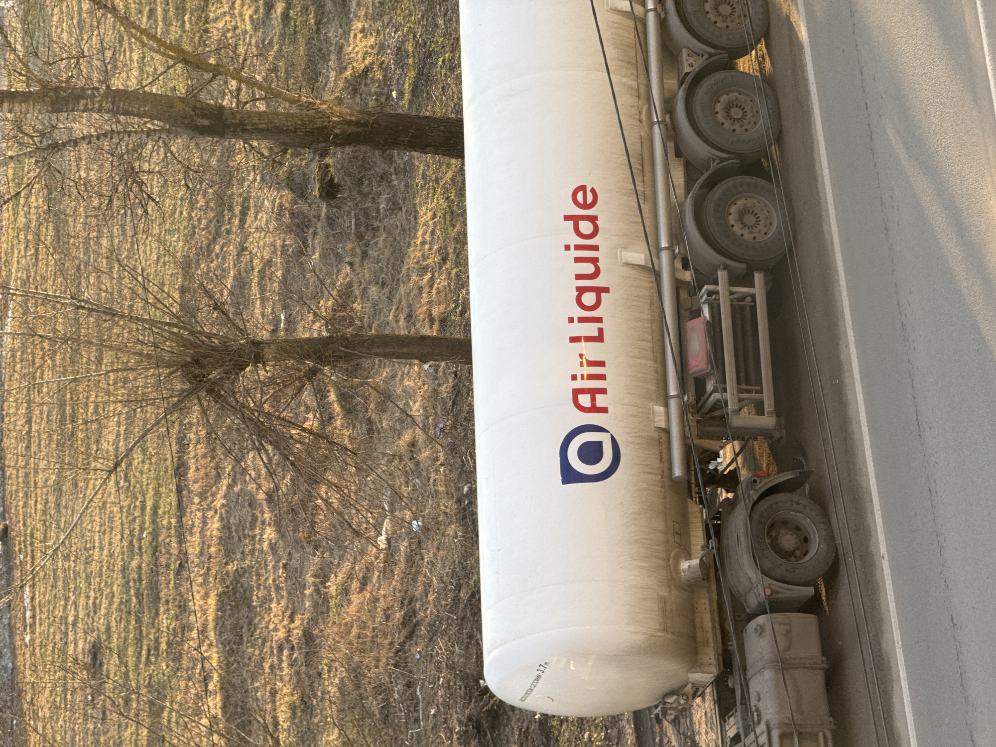 A white Air Liquide tanker truck is parked on a grey asphalt road in Kladovitsy, Russia. The truck's tires are visible on the right side of the image. The background features a field with dry grass and bare trees. Sunlight illuminates the scene.
