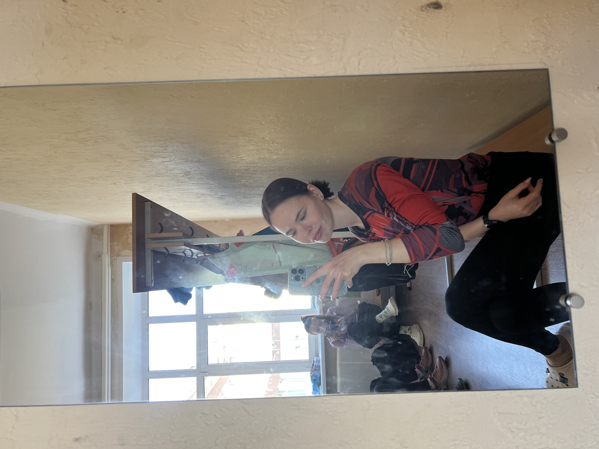 The image displays a mirror selfie of a young woman taken in an indoor setting, likely during the daytime. The woman, who has dark hair tied back, is wearing a red and black patterned long-sleeved top and black leggings. She is holding a smartphone with a light blue, patterned case to capture her reflection, and exhibits a slight smile. A watch or fitness tracker is visible on her left wrist, and she wears a ring on her right hand.
Reflected in the mirror is a brightly lit room with a large window composed of multiple panes, revealing bright daylight outside and what appears to be other buildings. To the left of the woman's head, a wooden rack or shelving unit with hooks is visible. The floor, seen in the reflection, is made of wood or laminate, with various shoes, including a pair of light-colored Crocs adorned with charms, scattered on it. Further in the background, a second figure—possibly another person or a deeper reflection—is visible near the window, seemingly sitting or crouching and also looking at a phone.
The walls surrounding the mirror are a light, textured beige or cream color. The mirror itself is mounted with visible silver standoffs or screws. Faint text, including the words "Krasnodar" and "Sabina," can be discerned on the wall above the mirror. The bright sunlight suggests a clear, sunny day.