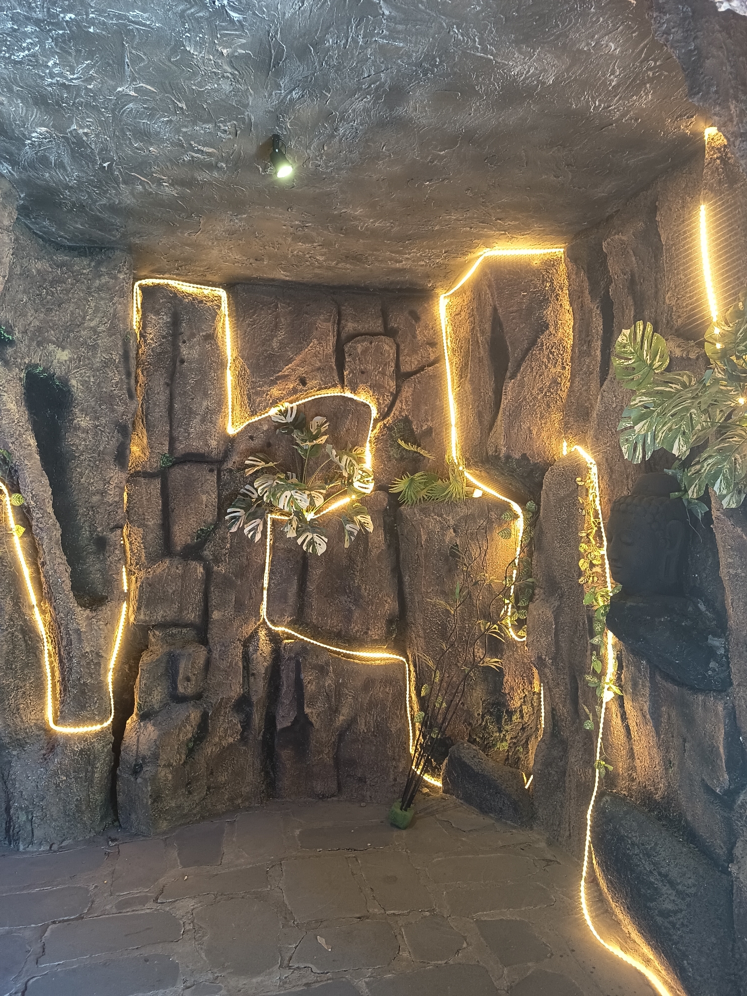 The image shows a dimly lit, cave-like interior with artificial rock formations and decorative lighting. The walls and ceiling are textured to resemble rough stone, with a rough, wavy pattern on the ceiling. Yellow LED strip lights are embedded along the edges of the rock formations, creating an illuminated path that outlines various sections of the walls.
Several artificial plants are placed within the scene. One prominent plant with variegated leaves is situated on a rock ledge. Another is a cluster of thin, dark stems in a green pot, placed on the floor in the lower right quadrant. Some climbing vines are also visible, adding to the natural, cave-like ambiance.
The floor is paved with large, irregularly shaped stone tiles, giving the appearance of an ancient pathway. A single, small spotlight fixture is visible on the ceiling. No people are present in the image.
The overall impression is that of an indoor decorative space designed to mimic a natural grotto or cave, likely a feature within a larger establishment. This scene is located in the city of Sawangan, Indonesia.