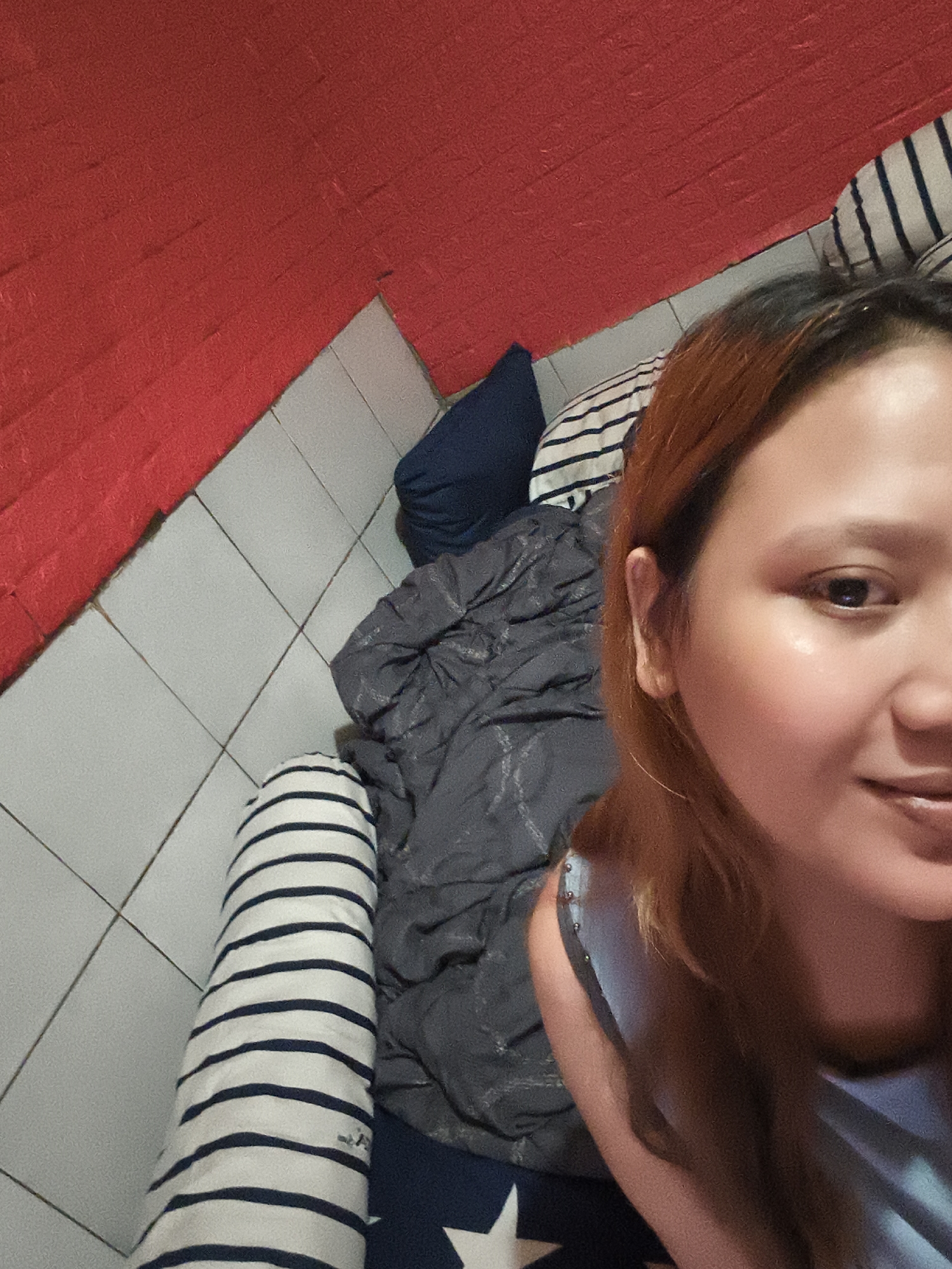 A selfie-style photograph taken indoors in Depok, Indonesia, shows a fair-skinned woman with reddish-brown hair looking at the camera with a slight smile. She is positioned on what appears to be a bed covered with striped cushions/pillows and a dark gray comforter. A blue pillow is also present. Behind the bed, the wall is tiled white with a red section above the tiling.