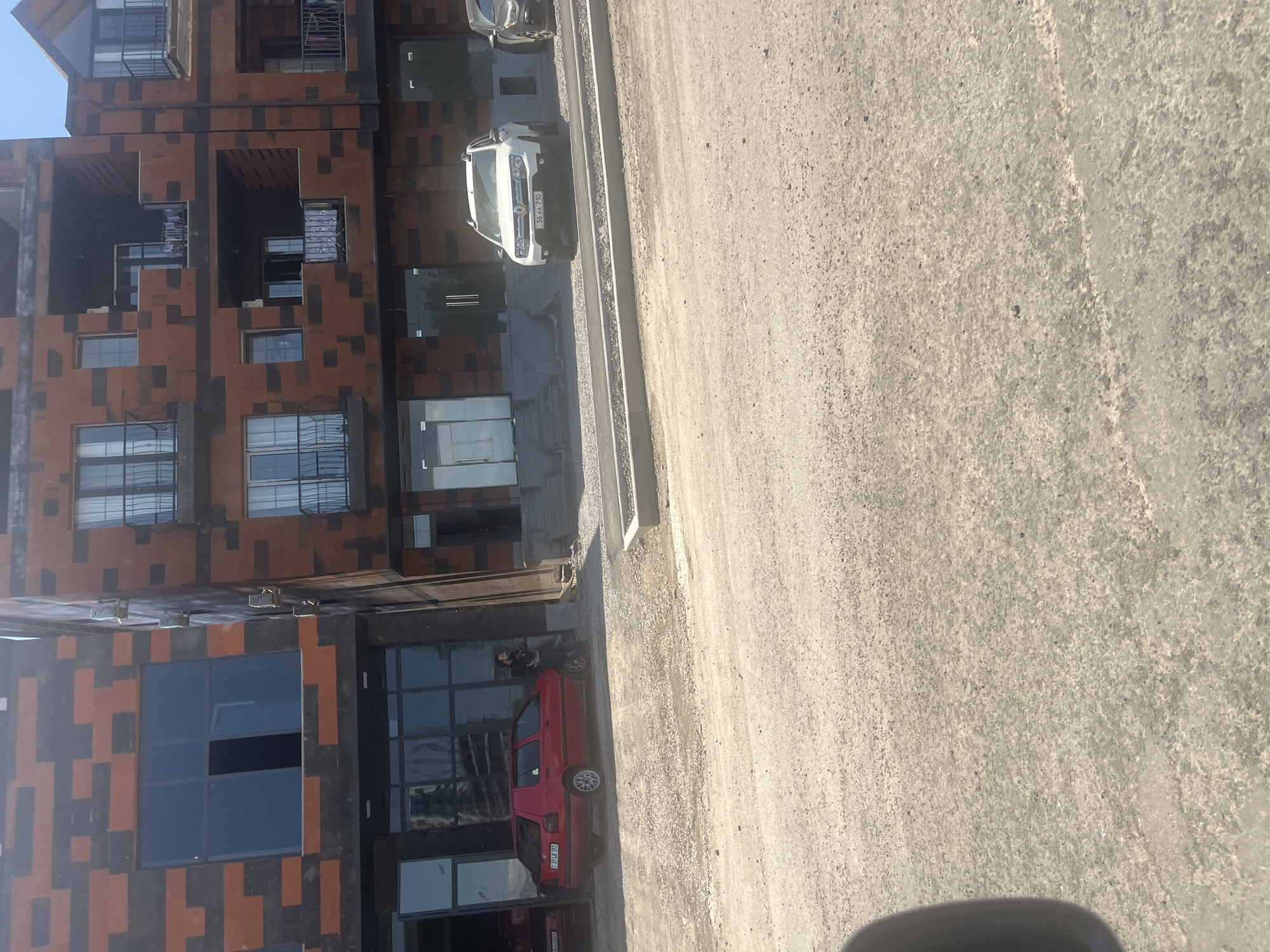 The image displays an outdoor scene in Abovyan, Armenia, characterized by modern architecture and an unpaved road under bright daylight.
On the left side of the frame, several multi-story buildings stand prominently. These structures feature a distinctive facade pattern composed of interlocking brown and black rectangular shapes, giving them a contemporary, pixelated appearance. The buildings have numerous windows, including large, reflective panels at ground level that suggest commercial establishments, as well as smaller windows and balconies on upper floors.
Adjacent to the buildings is a wide, unpaved area covered in light-colored dirt and gravel, serving as a road or parking space. Two cars are visible on this surface. In the foreground, closer to the viewer, a red sedan is parked next to the building. A person, dressed in dark clothing, is standing beside the driver's side of this red car, looking towards it. Further down the road, a white SUV is parked parallel to the buildings.
The time of day is bright and sunny, indicated by the clear blue sky visible at the top of the buildings and the strong shadows cast by the structures and vehicles. The weather appears clear and pleasant.
Visible text includes the license plates on both cars. The white SUV's license plate reads "36 AA 710". The red sedan's license plate reads "08 QC 700".