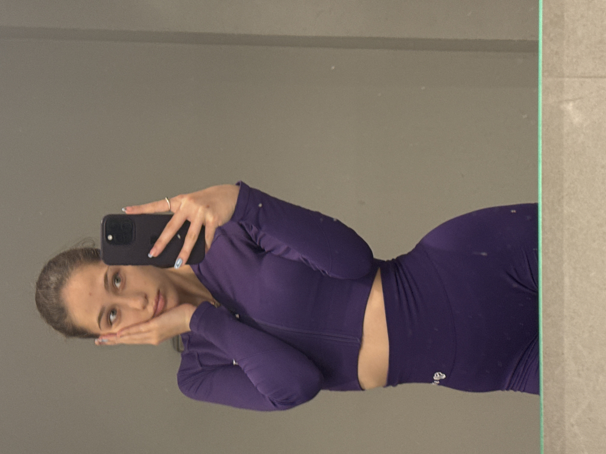A young woman is seen in a selfie taken in front of a mirror. She is wearing a matching purple athletic set, consisting of a long-sleeved crop top with a zipper and leggings. Her hair is pulled back, and she is holding a black smartphone with her left hand, resting her face on both hands. The background is a neutral gray wall, likely indoor. The location is unknown, but the prompt suggests it is in Sofia, Bulgaria.