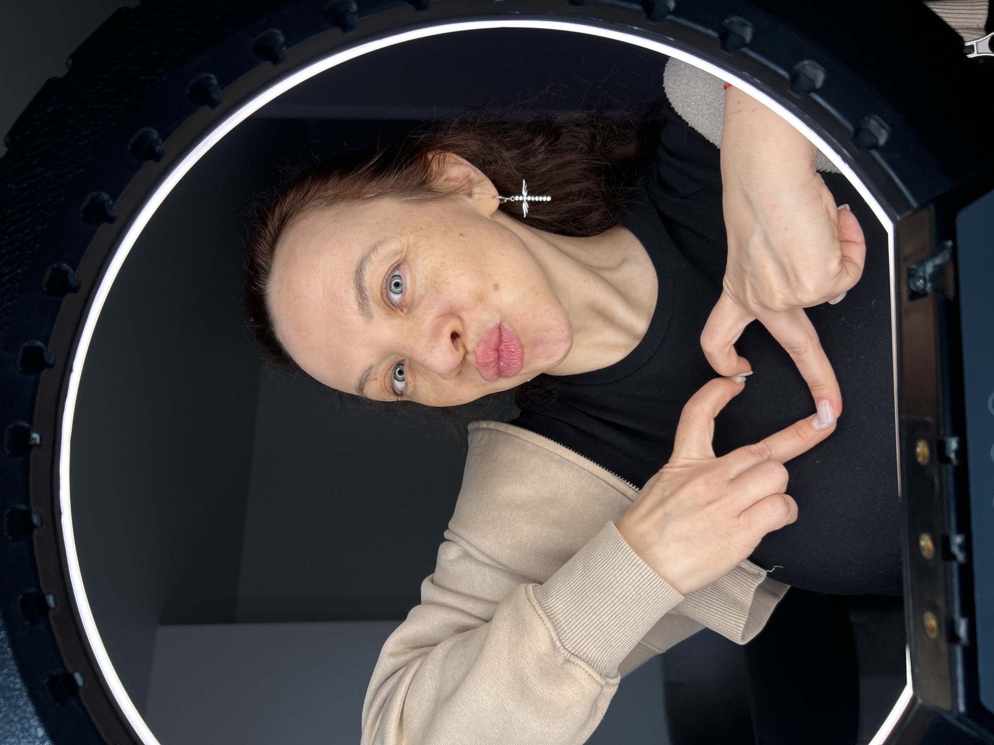 A woman with fair skin and blue eyes is looking at the camera while making a kissing face and a heart shape with her hands. She has brown hair and is wearing a black shirt and a beige hooded sweatshirt. She is wearing a silver cross earring in her left ear. The woman is positioned inside a ring light. The background is dark. The location of the image is Chaikovsky, Russia.