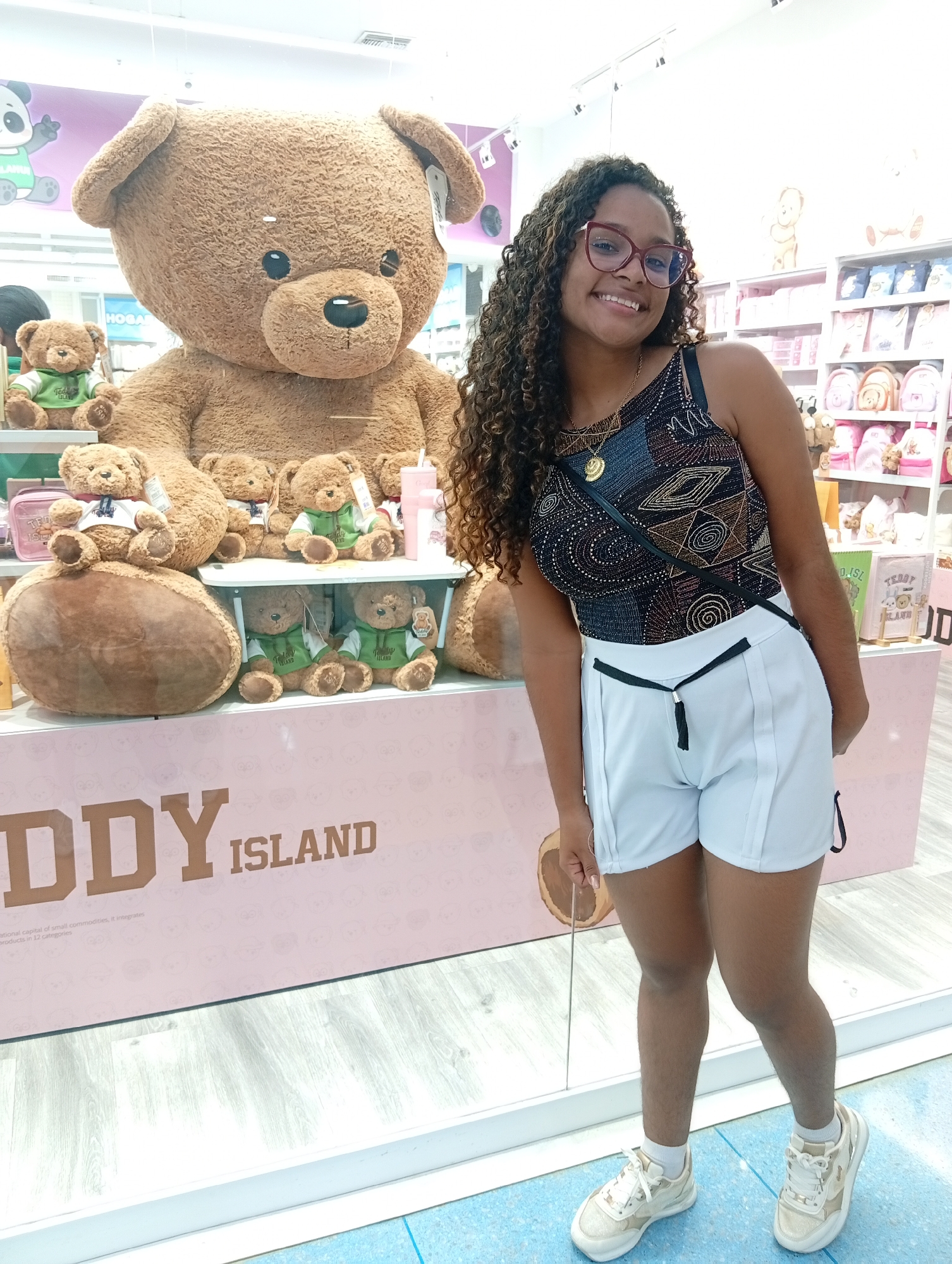The photograph shows a smiling woman with curly hair and glasses standing in front of a "Teddy Island" display in Porlamar, Venezuela. She is wearing a patterned one-piece swimsuit, white shorts with a black belt, white socks, and white sneakers. A black purse strap is visible across her torso.
Behind her is a large pink and white display with the words "TEDDY ISLAND" written in bold letters. On top of the display are several teddy bears, including one exceptionally large brown bear. Smaller teddy bears are arranged around it, some wearing green sweaters and others white shirts with bows.
In the background, shelves stocked with merchandise, possibly related to teddy bears or children's products, are visible. The floor in front of the display has a light blue and white pattern.
