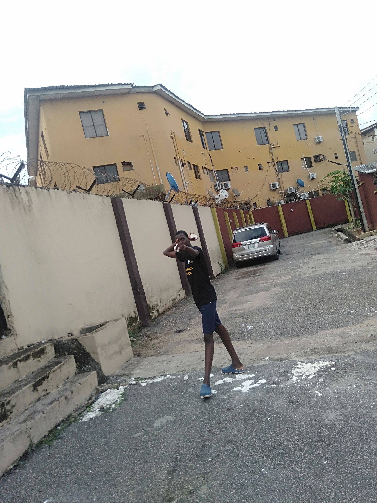 A medium shot shows a young person standing in a street in Shangisha, Nigeria. The person is dark-skinned, wearing a black t-shirt and blue shorts with blue slip-on sandals. The person is holding their hand to their head, making a triangular shape with their fingers and thumbs. Behind the person is a beige-colored wall topped with barbed wire and a beige-colored three-story building with windows, air conditioning units, and satellite dishes. A silver minivan is parked between the wall and a brown painted metal fence. The street is paved with asphalt. The sky is overcast.