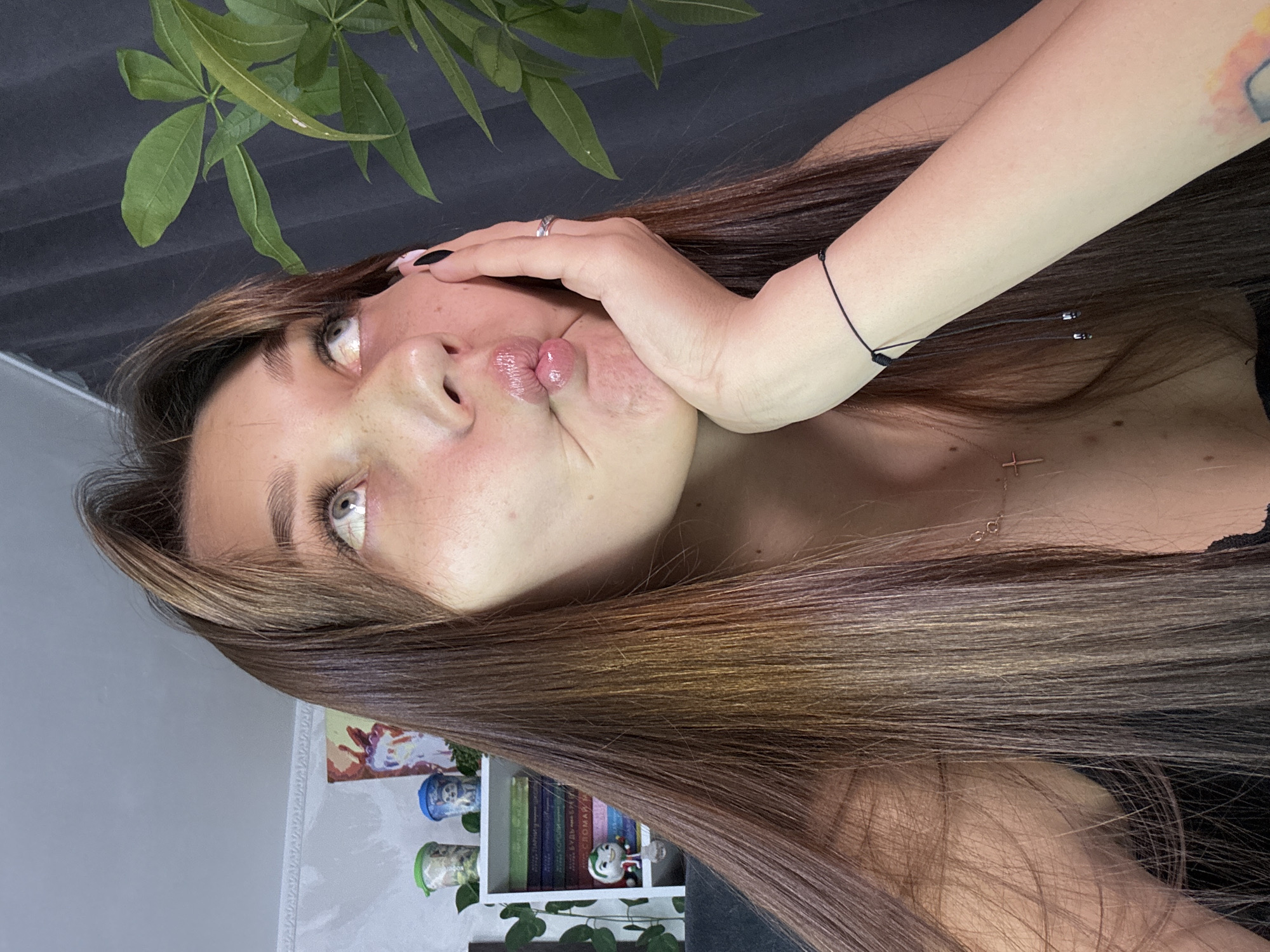 The image shows a young woman with long, dark brown hair, styled with bangs. She is looking up and to her right, with her lips puckered and her right hand resting on her cheek. She is wearing a black top, a delicate necklace with a cross pendant, and a thin black bracelet on her right wrist. Her right hand has black nail polish, and she is wearing a ring on her left ring finger. The background is blurred but suggests an indoor setting with shelves holding books and decorative items. To the right of the woman, there is a large green plant with broad leaves. There is no information in the image that would allow for the determination of its location in Novokuznetsk, Russia.