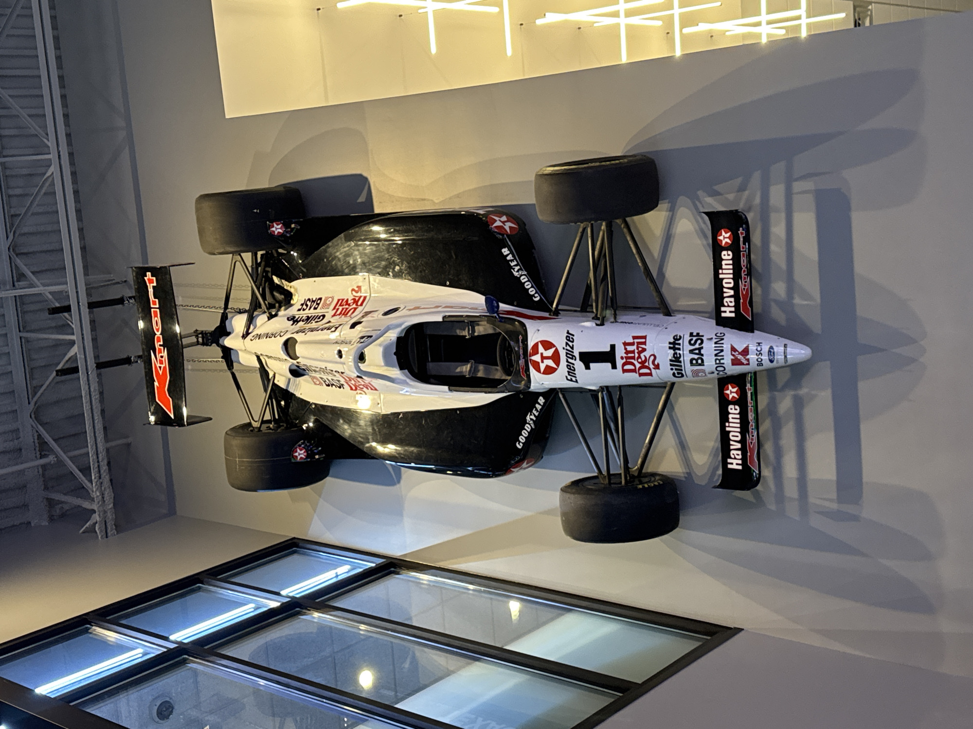 Here is a forensic description of the image:
The image shows a race car, specifically an IndyCar, mounted on a wall, seemingly as part of an exhibit. The car is predominantly white with black accents and various sponsor logos, including "Energizer," "Dirt Devil," "Gillette," "BASF," and "Havoline." The number "1" is prominently displayed on the side of the car. The car has four large racing tires, and the rear wing bears the "Havoline" logo. The exhibit is well-lit, and the car is displayed against a light gray wall. Below the car, there is a section of glass panels, possibly part of the building's architecture or another display element. The upper section reveals neon lighting. The lighting in the location is artificial, highlighting the vehicle on the wall.
I cannot provide the specific location in Schaumburg, United States of America.
