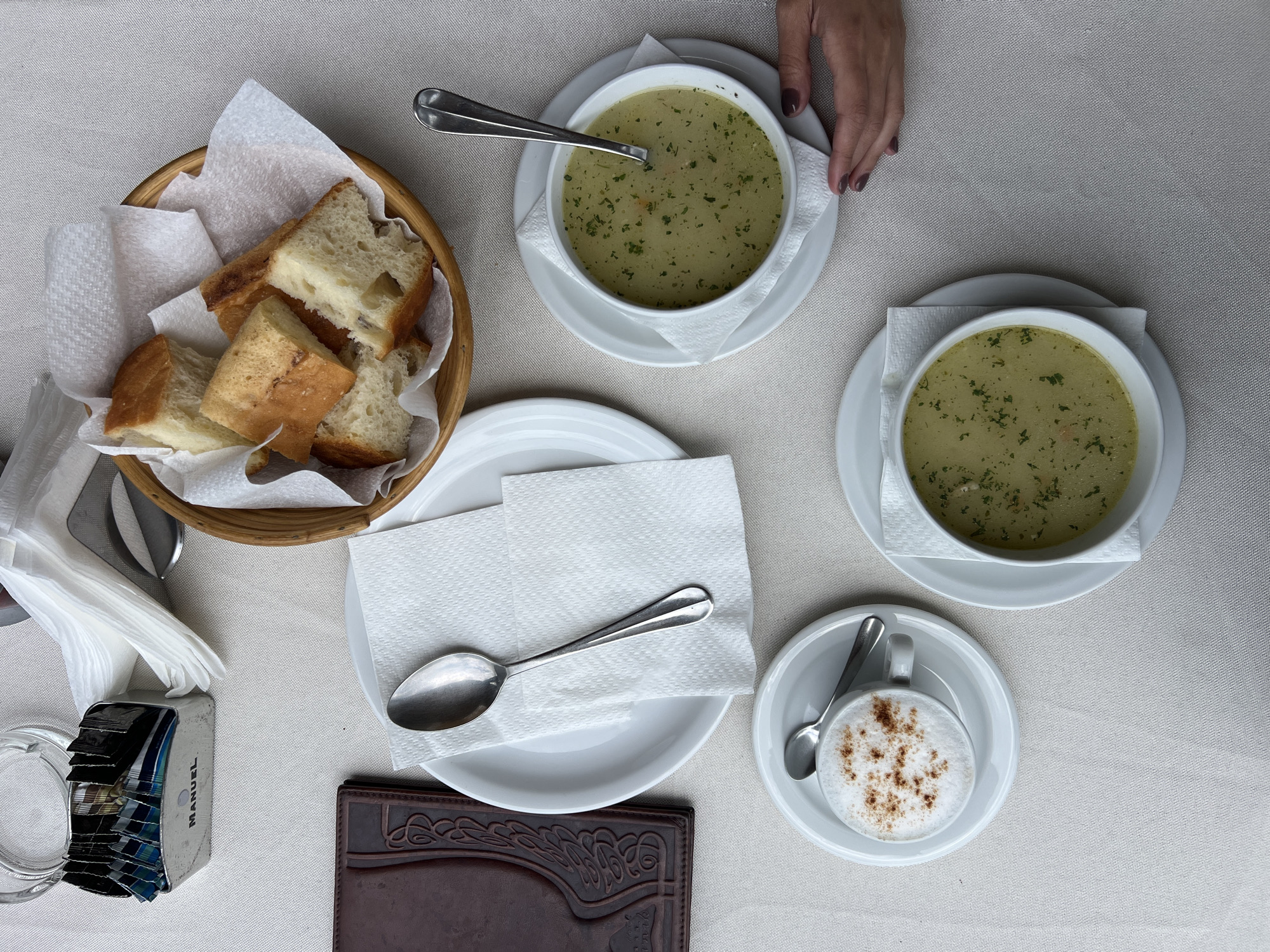 An overhead view captures a dining table set for a meal, likely in a restaurant or cafe in Šavnik, Montenegro. The table is covered with a light-colored, textured tablecloth.
The primary subjects are two bowls of soup, each served in a white bowl on a white saucer. The soup is light green or yellowish, garnished with chopped green herbs, possibly parsley, and appears to contain small vegetable pieces. A spoon rests in one of the soup bowls. To the left, a rustic woven basket holds several pieces of white bread, some with a golden-brown crust, lined with white paper napkins.
A person's hand, with dark-colored nail polish, is visible at the top right of the frame, resting near one of the soup bowls, suggesting the presence of a diner. In the lower right corner, a small white cup containing a hot beverage, likely a cappuccino or coffee, is topped with frothed milk and sprinkled with a brown powder, such as cinnamon or cocoa. It sits on a matching saucer with a small spoon.
Other items on the table include a stack of folded white paper napkins, a silver spoon resting on a white plate, and a clear glass, possibly an ashtray or beverage glass. A rectangular dispenser with various packets, possibly sugar or creamers, is also visible, bearing the brand name "MANUEL" on its side. A dark brown wallet or menu holder with an embossed pattern is positioned towards the bottom center. The scene suggests a casual meal for two.