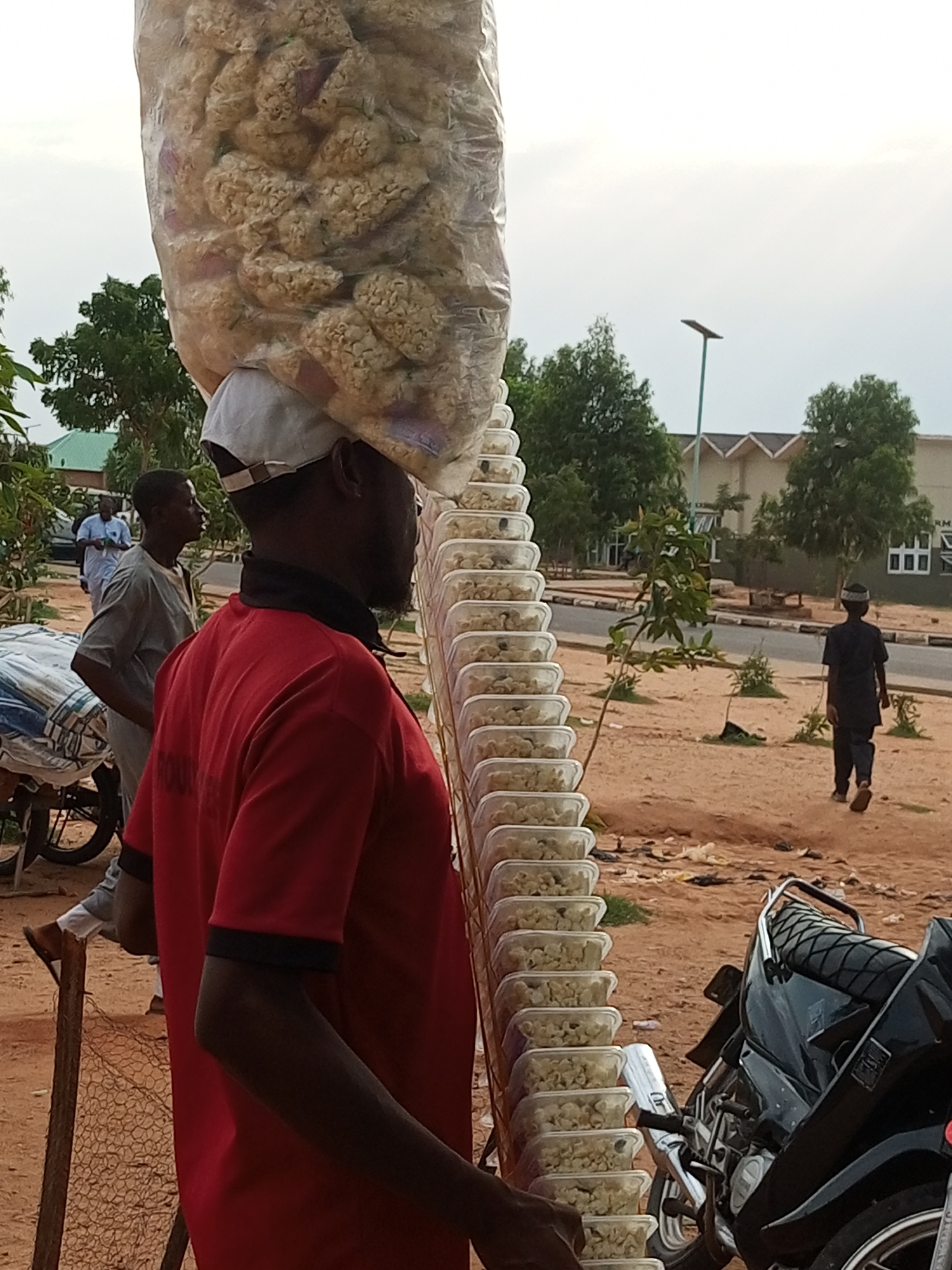 The image captures a street scene in Gombe, Nigeria, with a focus on a man carrying a large bag of what appears to be snacks on his head. He is wearing a red t-shirt and a white cap. In front of him, a stack of clear plastic containers, also filled with snacks, is arranged in a vertical line. To his left, a motorcycle is parked, and a man wearing a grey tunic stands behind it. In the background, another person is seen walking away, dressed in dark clothing. The environment appears to be an outdoor setting with sandy ground, trees, and buildings in the distance, suggesting an urban or semi-urban area. The sky is overcast.