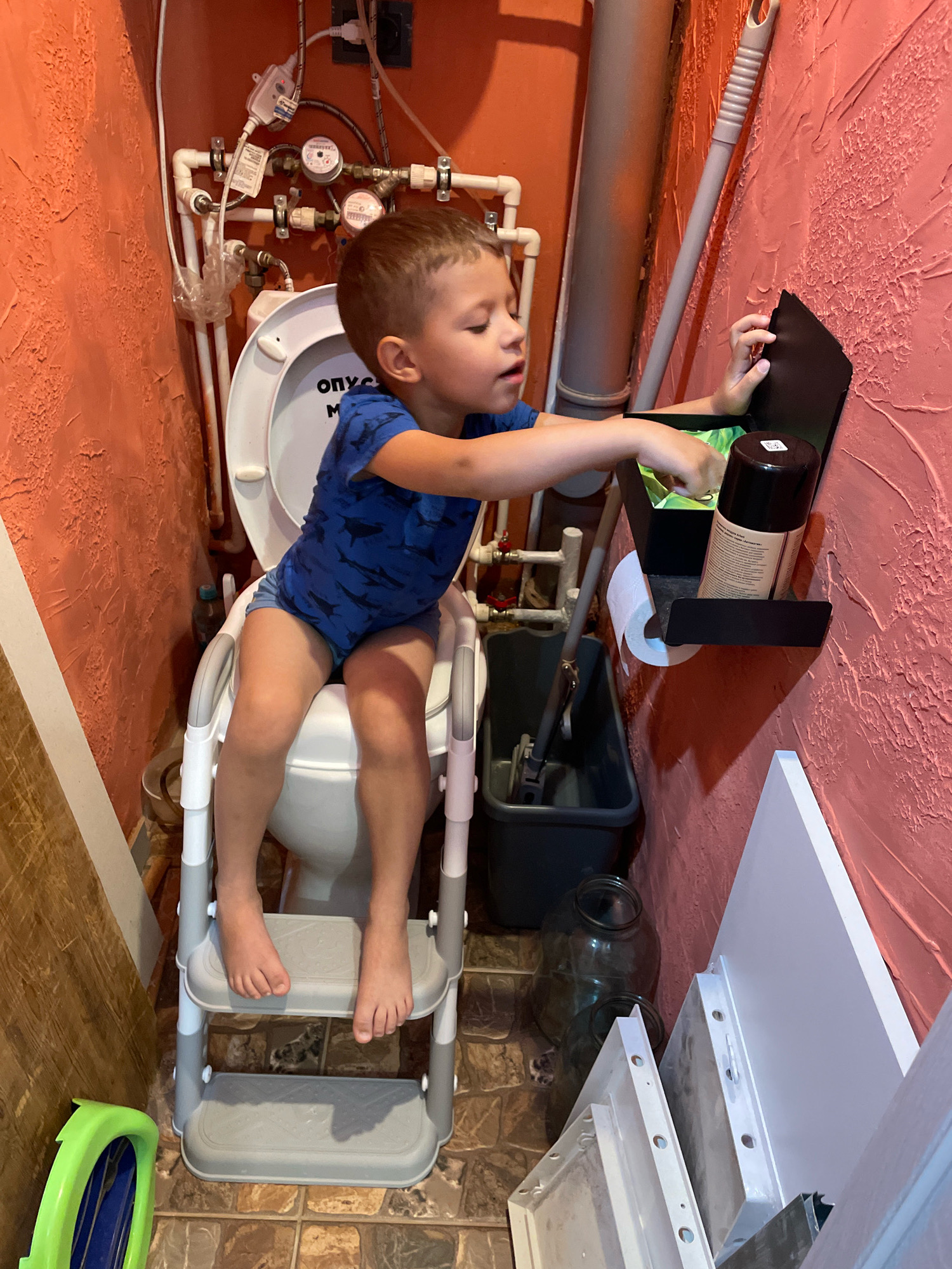 A young boy, appearing to be around 3-4 years old, is sitting on a portable potty training seat placed on top of a toilet. He is wearing a blue t-shirt with a shark print and blue shorts. His feet are resting on a two-step stool. The boy's right hand is reaching into a black shelf mounted on the pink, textured wall. On the shelf are two spray cans, one with a white label and a black cap, and another green item in a bag. A roll of toilet paper is also on the shelf. The environment appears to be a small bathroom, with plumbing fixtures and pipes visible behind the toilet. A mop and bucket are on the floor to the right of the toilet. There are large glass jars and some white boards stacked near the wall to the right. The floor is tiled. The image was captured in the city of Arkhangelsk, Russia.